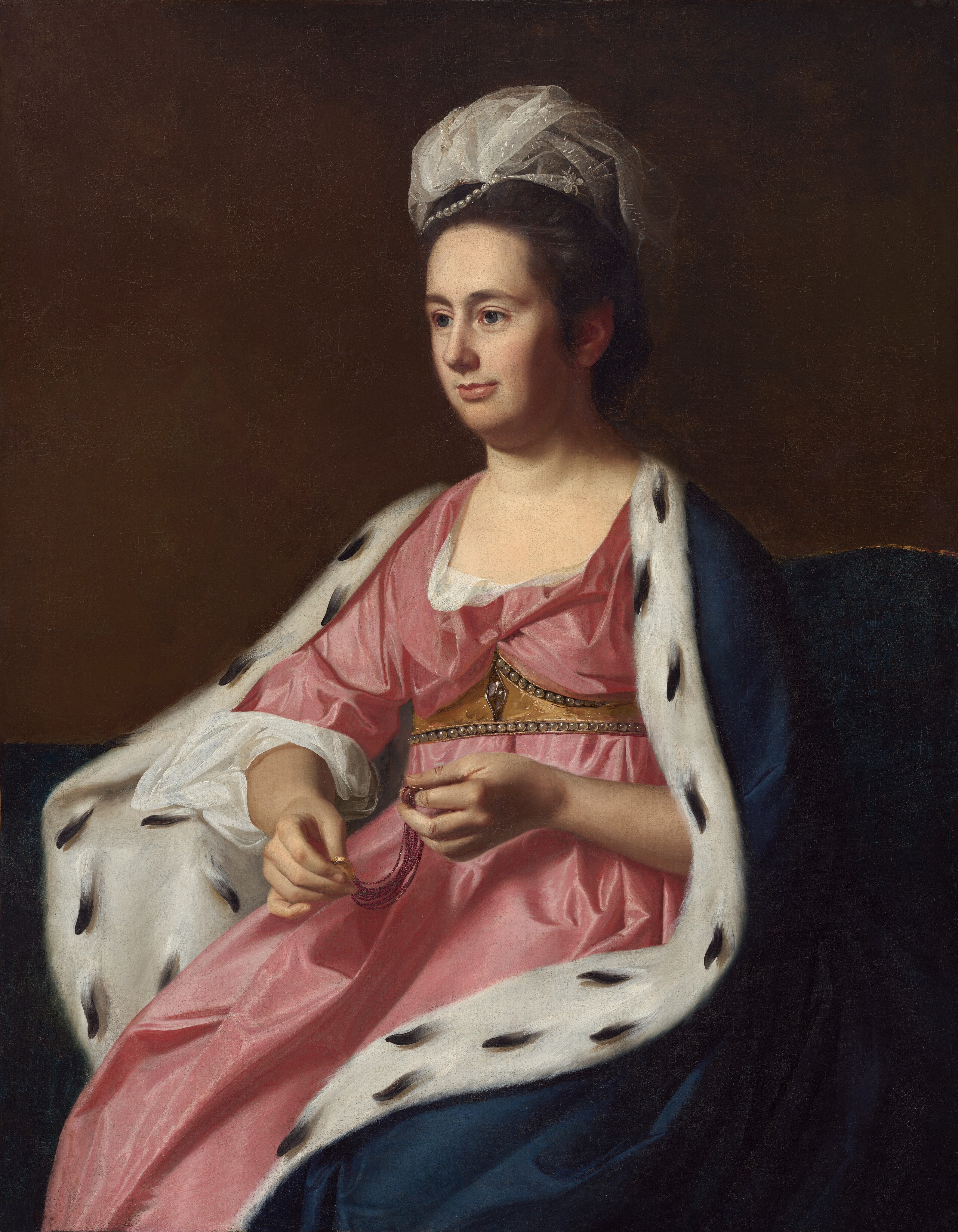 Shown from the knees up, a pale-skinned woman looking off to the left from her seat on a divan wears a carnation-pink gown and a fur-lined cloak in this vertical portrait painting. Her body is angled to our left, and she looks into the distance with close-set, large, gray-blue eyes. She has dark brows, a straight nose, a full lower lip, and a slight double chin in her long, oval face. Pewter-gray hair is brushed back from her high forehead and bound up under a loose swath of lace and pearls. Her loose gown is gathered under a gold, pearl-lined belt that follows the shape of her breasts along the top edge and is straight across the bottom edge. Tufts of snow-white cloth are pulled out at the neckline and at the elbow. The skirt falls loosely under the cloak, which is marine blue on the outside. The white fur interior is speckled with black, suggesting ermine. The woman holds two ends of what might be a bracelet made up of at least ten strands of fine, purple beads. Each end has a gold fitting, perhaps clasps. Her navy-blue seat curves from behind her shoulders down and across the background under a coffee bean-brown background. The woman is lit starkly from the left, throwing her ear and the far side of her face, to our right, into shadow.