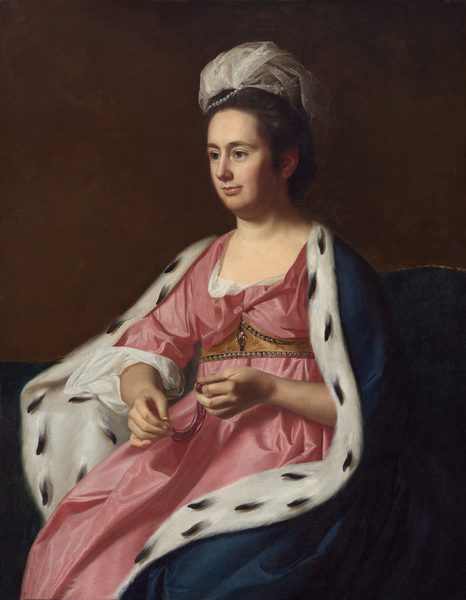 Shown from the knees up, a pale-skinned woman looking off to the left from her seat on a divan wears a carnation-pink gown and a fur-lined cloak in this vertical portrait painting. Her body is angled to our left, and she looks into the distance with close-set, large, gray-blue eyes. She has dark brows, a straight nose, a full lower lip, and a slight double chin in her long, oval face. Pewter-gray hair is brushed back from her high forehead and bound up under a loose swath of lace and pearls. Her loose gown is gathered under a gold, pearl-lined belt that follows the shape of her breasts along the top edge and is straight across the bottom edge. Tufts of snow-white cloth are pulled out at the neckline and at the elbow. The skirt falls loosely under the cloak, which is marine blue on the outside. The white fur interior is speckled with black, suggesting ermine. The woman holds two ends of what might be a bracelet made up of at least ten strands of fine, purple beads. Each end has a gold fitting, perhaps clasps. Her navy-blue seat curves from behind her shoulders down and across the background under a coffee bean-brown background. The woman is lit starkly from the left, throwing her ear and the far side of her face, to our right, into shadow.