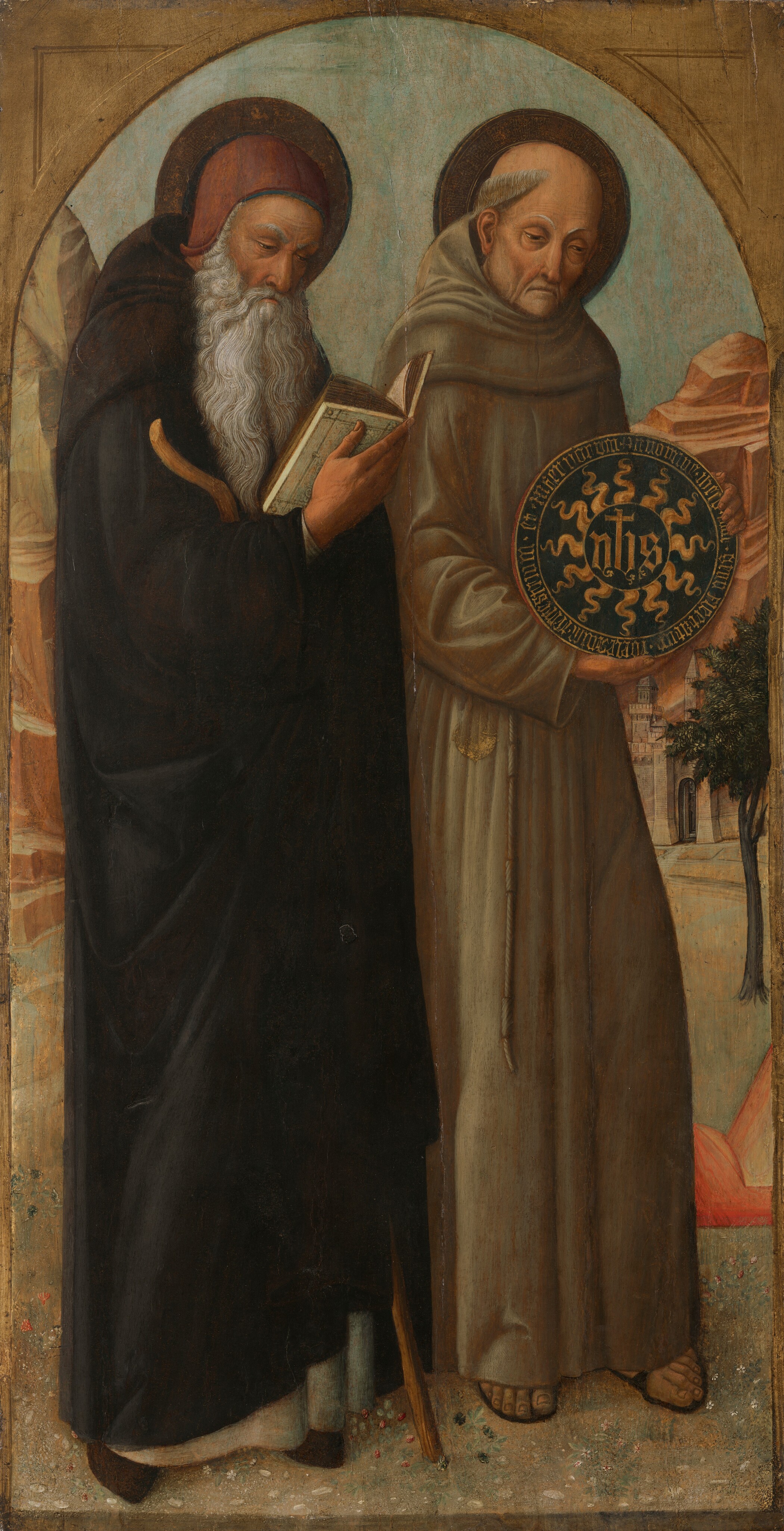 This painting shows two men standing side by side, both partially turned to the right. The man on the left is reading a book and holding a wooden staff. He has a long white beard and wears a black robe and a red cap. The man on the right has a round, balding head with traces of gray hair above his ear, and his face is gaunt. He wears a brown robe and holds a large circular object with a yellow sun-like shape on a black background, some letters at the center and more words around the rim. Both men have pale skin and dark brown halos around their heads. The background shows tall red and gray rocks, a distant gray building, and a thin tree with dark leaves, all under a blue sky. The painting is framed by a gray arch.