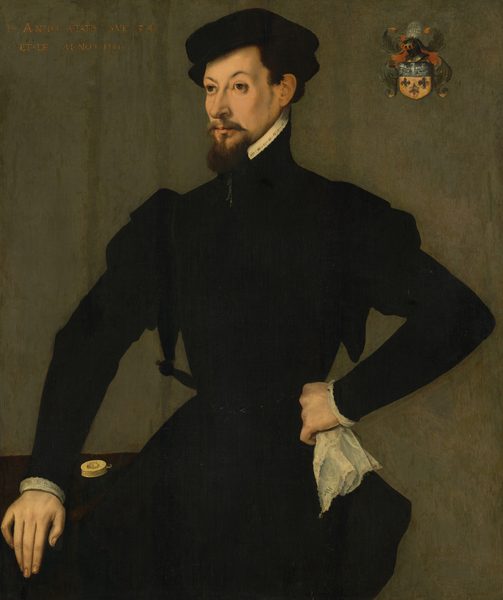 The portrait shows a man from the waist up. He is positioned at an angle with his left hand resting on a surface and his right hand on his hip, holding a white lace handkerchief. He has sharp facial features with a short dark beard and mustache, and his hair is styled neatly beneath a black cap. He is dressed in a black outfit with a high collar, and a small area of white shirt is visible at the wrist and collar. On the surface beside him rests a pocket watch. The background is plain and features a coat of arms in the upper right corner.