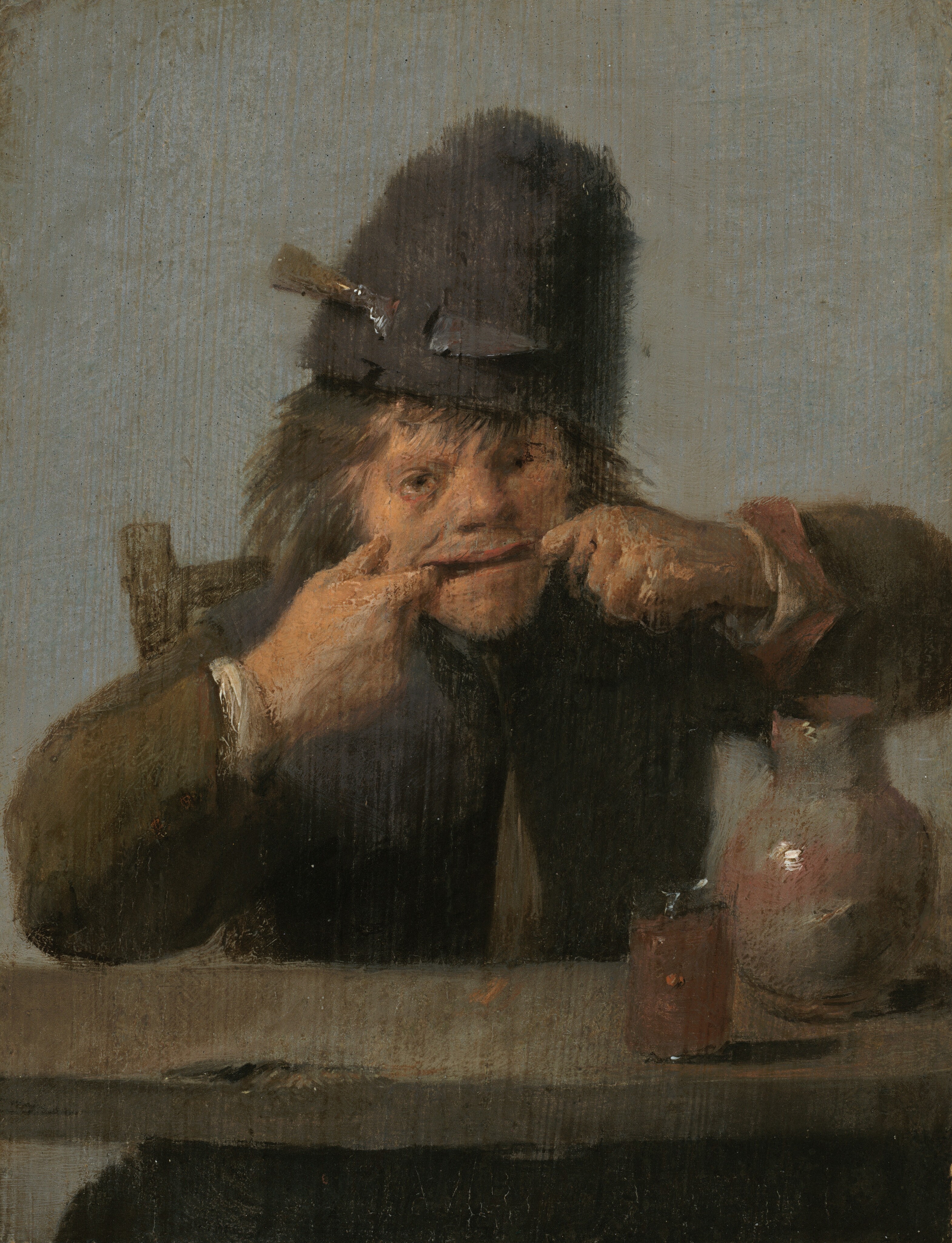 A young man with tanned, peachy skin sits at a table pulling his mouth wide with both hands in this vertical painting. On the far side of a wooden table, he faces us with elbows splayed wide. He has hooked the first finger of each hand into the corners of his mouth to stretch his open lips into a long, goofy line. He has wide-set eyes and a wide nose. His brown hair sticks out from under a tall, fuzzy brown hat, which has a knife secured by the blade in a loop to our left. The man’s iron-gray jacket has coffee-brown sleeves with the white of his shirt showing at the cuffs. A putty-gray jug accented with washes of rust-red and a glass filled with an amber liquid sit on the table before him. Soft light from the upper left gleams white on these smooth surfaces. A dark mass visible under the table is presumably the tops of his legs. The top right corner of his wooden chair is visible over his right shoulder, on our left, and he is set against a dove-gray background. The scene is loosely painted so brushstrokes are especially visible in the man’s face and hands.