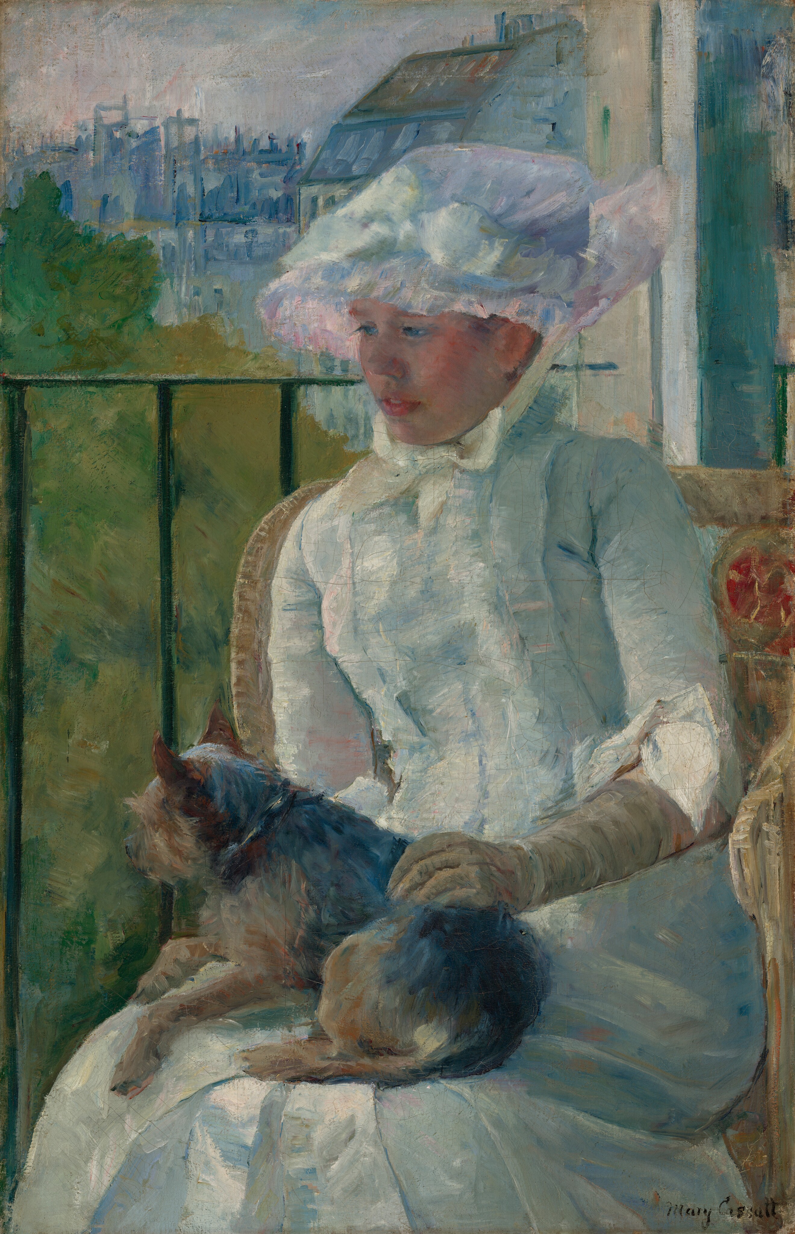 Shown from the knees up, a young woman wearing a white gown and bonnet sits in front of a balcony railing on a sunny day, holding a small dog on her lap in this vertical portrait painting. The scene is painted with visible brushstrokes, which are especially broad in the background and clothing. The woman sits with her body angled to our left, and she looks down in that direction with blue eyes. She has a short, snub nose, rosy cheeks, and her full, pink lips are parted. The woman’s bonnet is tied under her chin with a pale yellow bow. Her dress has a tightly-fitting bodice, elbow-length sleeves, and a long skirt. The bonnet and dress are painted with strokes of faint sky blue, petal pink, lavender purple, and mint green, with cream-white highlights and some darker cobalt-blue and apricot-peach strokes for shading. The dog on her lap lies with its head to our left, lifted as it looks out onto the landscape. The tan glove the woman wears on the arm we can see reaches her elbow, and she rests that hand on the dog’s back. Her right arm, to our left, is hidden below the elbow by the dog. The animal has charcoal-gray fur along its back and tan fur on the legs and shaggy face. The woman sits in a wicker armchair with a crimson-red medallion next to her left shoulder, to our right. The black balcony railing spans the width of the painting at the level of her nose, and has vertical bars. In the landscape beyond, trees to the right are painted loosely with sage green and mustard yellow. Buildings in the distance fill the top quarter of the composition, and are loosely painted with strokes of slate and aquamarine blue. The sky above is streaked with pale blush pink and ice blue. The artist signed the painting in the lower right corner, “Mary Cassatt.”