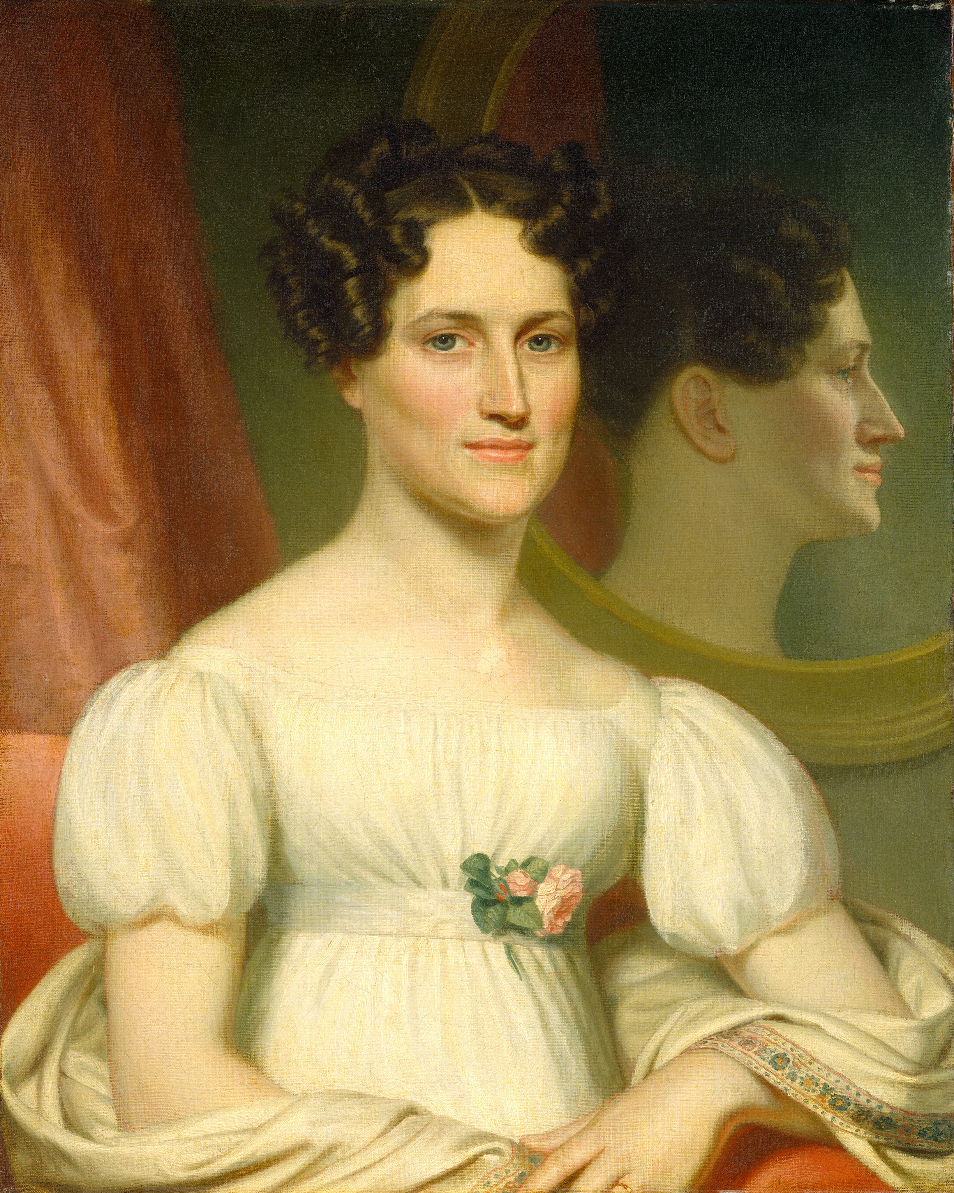 The image shows a portrait of a person from the mid-torso upwards. The person is seated, facing forward, with a mirror behind reflecting the side of her face in profile. She has fair skin, dark curly hair, and is wearing a light-colored dress with puffed short sleeves and a floral element at the bust. A patterned fabric is draped over her right arm as she sits with her fingers resting on her lap. The background is simple with a dark greenish hue, partially showing a large oval mirror and a reddish curtain on the left.