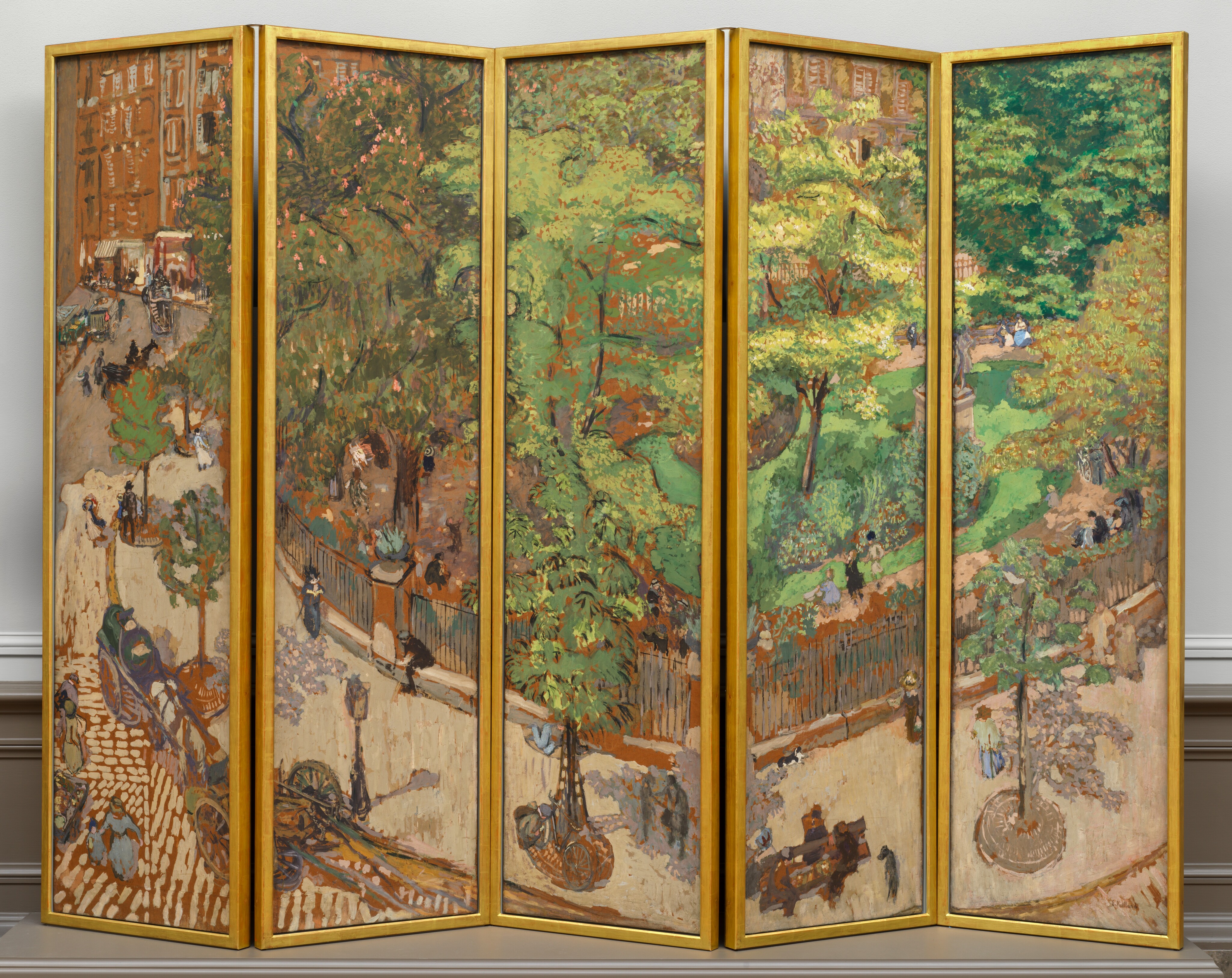 A screen made up of five tall, rectangular panels, set side by side and each surrounded by a gold frame, is painted as a single scene showing a tree-lined sidewalk curving around a park in a city. The scene is loosely painted with short, rounded brushstrokes. The top two-thirds to three-quarters of most of the panels are filled with the lime and olive-green leaves of the trees that line the sidewalk and park. In the leftmost panel, the sidewalk and road lead back to a row of caramel-brown building façades. The sidewalk is pale taupe, and the street is painted with dashes of the same taupe against terracotta brown, suggesting cobblestones. Spindly trees are spaced in a row along the sidewalk in round holes covered with smoke-gray metal grates. A black fence, painted with thin, sometimes broken black lines encloses the park beyond, which has a path around plantings and the vivid green lawn. Touches of pink on a sage-green tree to our left in the park suggest flowers. A gray statue on a high plinth is partially lost in the break between the two rightmost panels. Men, women, and children, painted with a few strokes of black, gray, or marine or periwinkle blue, walk along the sidewalk and the garden path, or sit at the base of the fence or on benches spaced along the sidewalk. The women seem to wear long dresses and the men dark clothing and hats. Two carriages are pulled up on the street near a lamp post alongside the sidewalk near the lower left. In the leftmost panel, horse-drawn carriages move along the road leading back to the buildings, and more people seem to be gathered on the sidewalk near the left edge of the panel in the distance. The artist signed the work with brown paint in the lower right corner: “E. Vuillard.” The panels of the screen have been set up so the panels rest on a platform or on the floor in a shallow zig-zag pattern, in a room with an off white wall and bisque-brown molding along the floor.