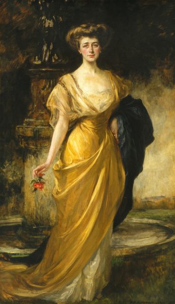 This painting shows a woman in a full-body portrait. She is standing with her body turned slightly to the right, looking out at us. The woman has pale skin, rosy cheeks, dark eyebrows, deep-set eyes, and red lips in a slight smile. Her dark hair is styled in an intricate updo. She wears a golden-yellow gown with a low neck, puffed, sheer sleeves, and a skirt that drapes down her legs and trails behind her. She holds a red flower in her right hand, and a dark wrap or shawl is draped over her left arm, her left hand on her hip. A gold ring is visible on her left hand, and she has round golden earrings. The background is dark, with what looks like a sculpture or fountain behind her on the left. The rest of the background is in tones of brown, green, cream, and pale yellow.