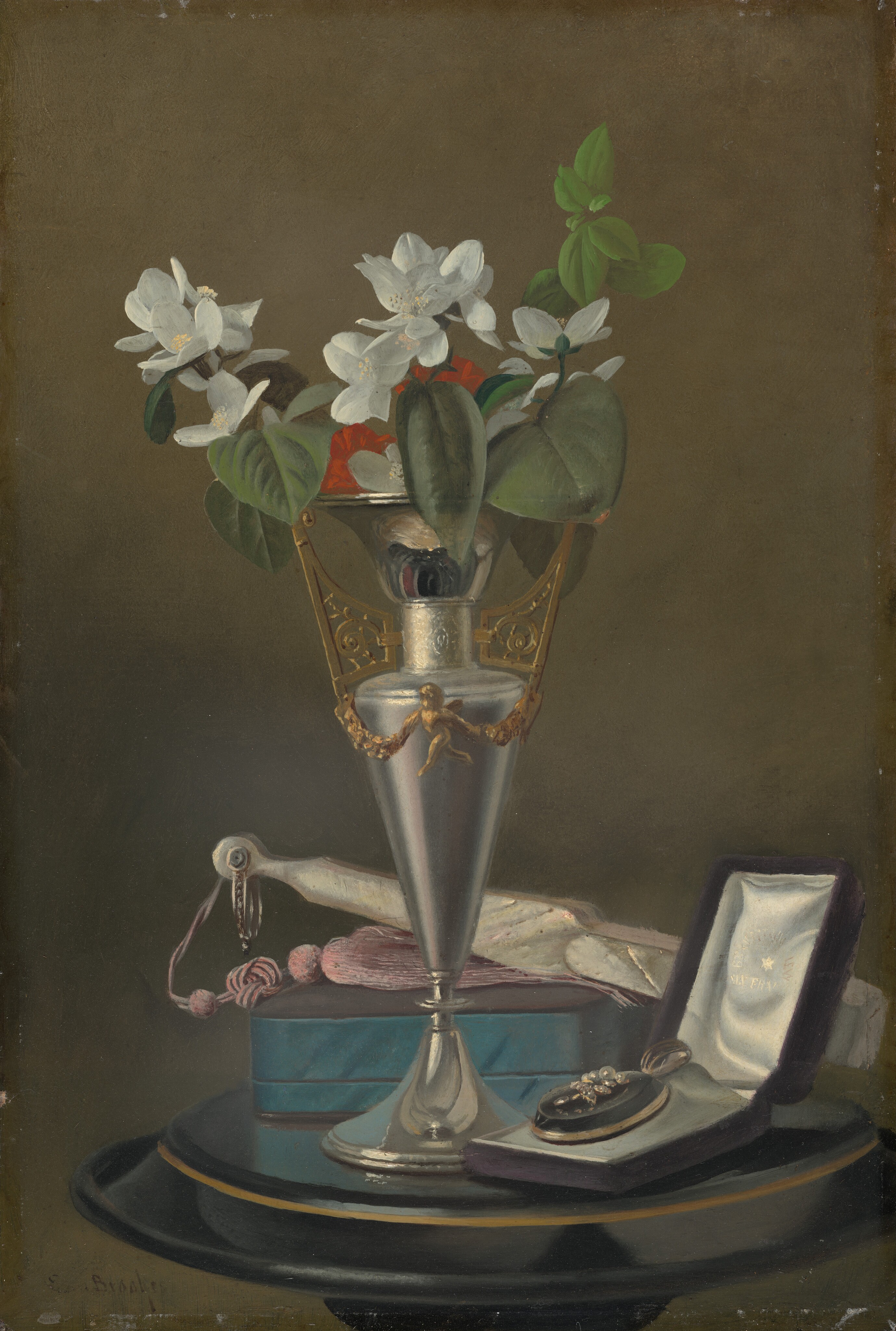 A tall silver vase with gold details holds a bouquet of white flowers with green leaves and red blossoms. The color palette includes metallic silver, gold, white, green, red, and teal. Positioned on the table, the vase stands centrally, with a blue box behind it to the left and a jewelry box with a pearl and flower ring design slightly to the right. There is also a piece of fabric with a tassel and small scissors lying flat behind the vase.