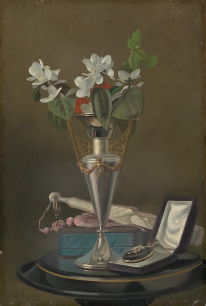 A tall silver vase with gold details holds a bouquet of white flowers with green leaves and red blossoms. The color palette includes metallic silver, gold, white, green, red, and teal. Positioned on the table, the vase stands centrally, with a blue box behind it to the left and a jewelry box with a pearl and flower ring design slightly to the right. There is also a piece of fabric with a tassel and small scissors lying flat behind the vase.
