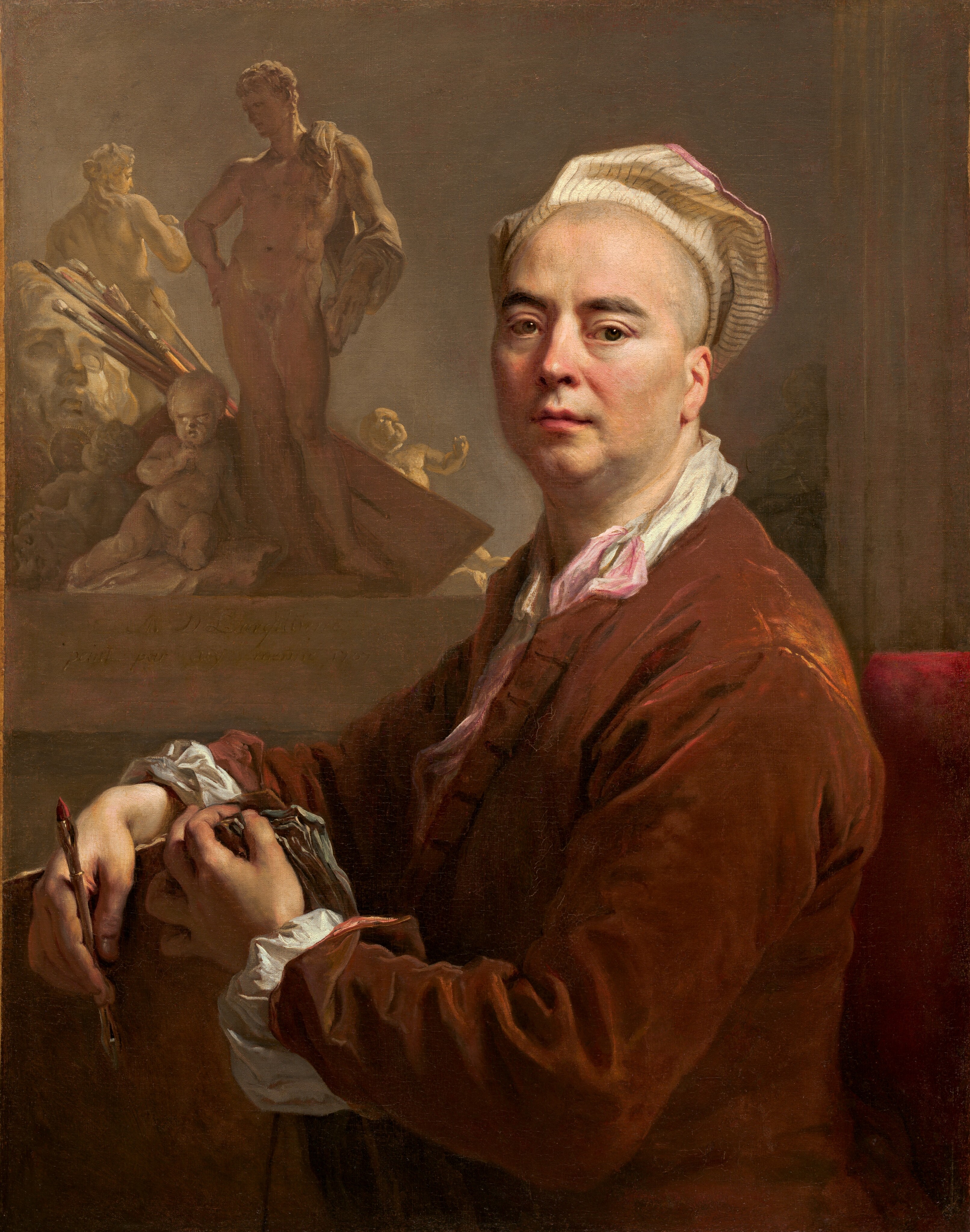 The image shows a portrait of a man visible from the waist up, seated in a red cushioned chair, his body turned to the left and his face turned to us. His facial features include pale skin, dark eyebrows, slightly sagging cheeks, and pink lips in a neutral expression. His hair seems to be shaved, and his head is mostly covered by a white, striped head covering that resembles a beret. He wears a reddish-brown coat with white ruffled sleeves, and underneath a white collared garment and a light pink ribbon peek out. In his hands, the man holds drawing instruments, including a pencil or pen and a stack of papers. Behind him on the left are several small tan and white sculptures, including a nude male figure, a cherub, and a woman's head. Beside these sculptures are several paint brushes and a flat surface that could be a painter's palette. The walls behind him are gray.