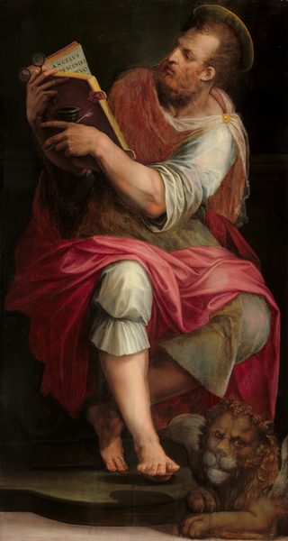 A pale-skinned, muscular man sitting and reading a book as a lion rests at his feet fills this vertical painting. The man’s knees are angled slightly to our right but his torso twists back to the left. His head follows that direction so he’s almost in profile as he looks at the book, which he holds up to our left. His brows are deeply furrowed, and he has a bumped, blade-like nose. He has short, copper-red hair and a squared beard. He wears a tunic and knee-lengths pants that shimmer in shades of topaz blue and pearly white under a carnation-pink cloak that drapes from his shoulders and across his thighs. A round, gold pin holds the cloth in place on his left shoulder, to our right. His lower legs and feet are bare. He rests the heel of one foot on a rounded plinth and lifts the toes so they come at us. The other knee bends deeply and the toes of that foot perch lightly behind the first leg. The man leans to our right to look at the open book, which he holds in both arms to the left. Visible text on the far page reads, “ANGELVS DESCENDIT EVO.” He braces a black inkpot and spindly white feather quill in one hand and holds a pair of pince-nez glasses with the other. The lion is small in comparison to the man and has a curly mane and translucent gray wings. The background is dark except a strip off-white floor along the bottom edge.