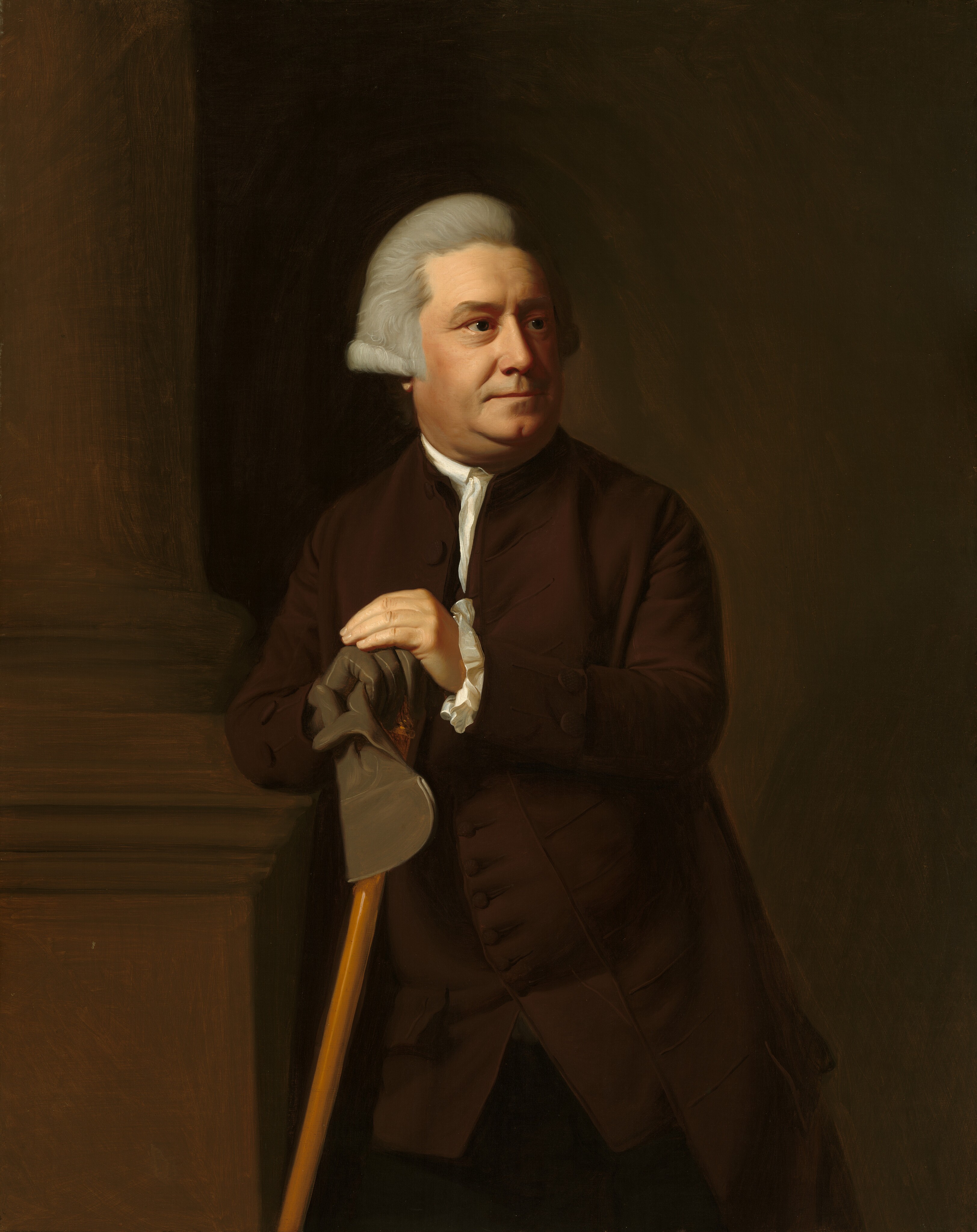 This painting shows a man from the knees up.  His body faces us, while his head is partially turned to the right. He has a round face with light skin and light gray hair combed back from his face and coming down to his ears. He has a straight nose, thin lips, and a double chin. He wears a dark brown coat with a white collar and cuffs and holds a light brown cane. One of his hands is wearing a dark gray glove, while the other is bare; the gloved hand also holds the other glove, along with the cane. The man is leaning against a large brown column that takes up the left side of the painting, and the wall behind him is dark brown.