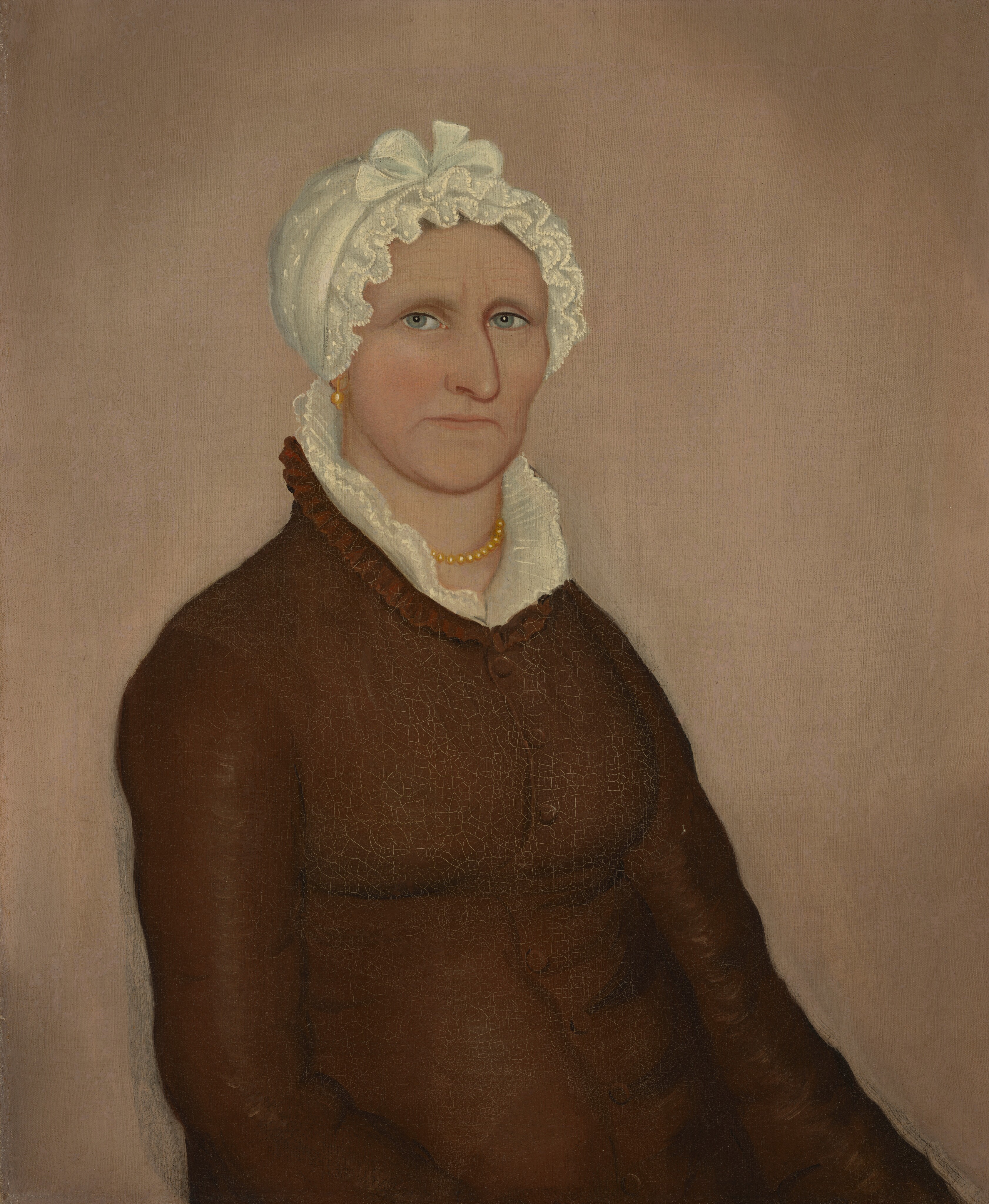 The image shows a portrait of a woman depicted from the chest up, seated and facing slightly to the left. Her facial features are defined with pronounced cheekbones, a firm jawline, and light-colored eyes set under well-defined eyebrows. Her hair is not visible as it is covered by a white cap adorned with lace and a bow on top. The woman wears a brown garment with a high collar and ruffled shirt underneath. Around her neck, she wears a string of yellow beads or pearls, and small earrings can be seen. The background is plain and neutral.