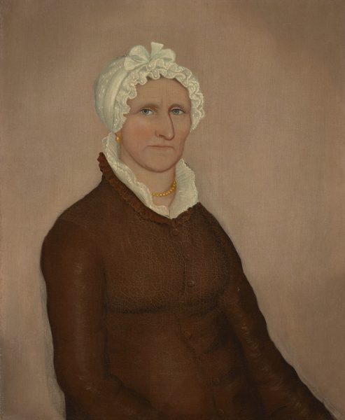 The image shows a portrait of a woman depicted from the chest up, seated and facing slightly to the left. Her facial features are defined with pronounced cheekbones, a firm jawline, and light-colored eyes set under well-defined eyebrows. Her hair is not visible as it is covered by a white cap adorned with lace and a bow on top. The woman wears a brown garment with a high collar and ruffled shirt underneath. Around her neck, she wears a string of yellow beads or pearls, and small earrings can be seen. The background is plain and neutral.