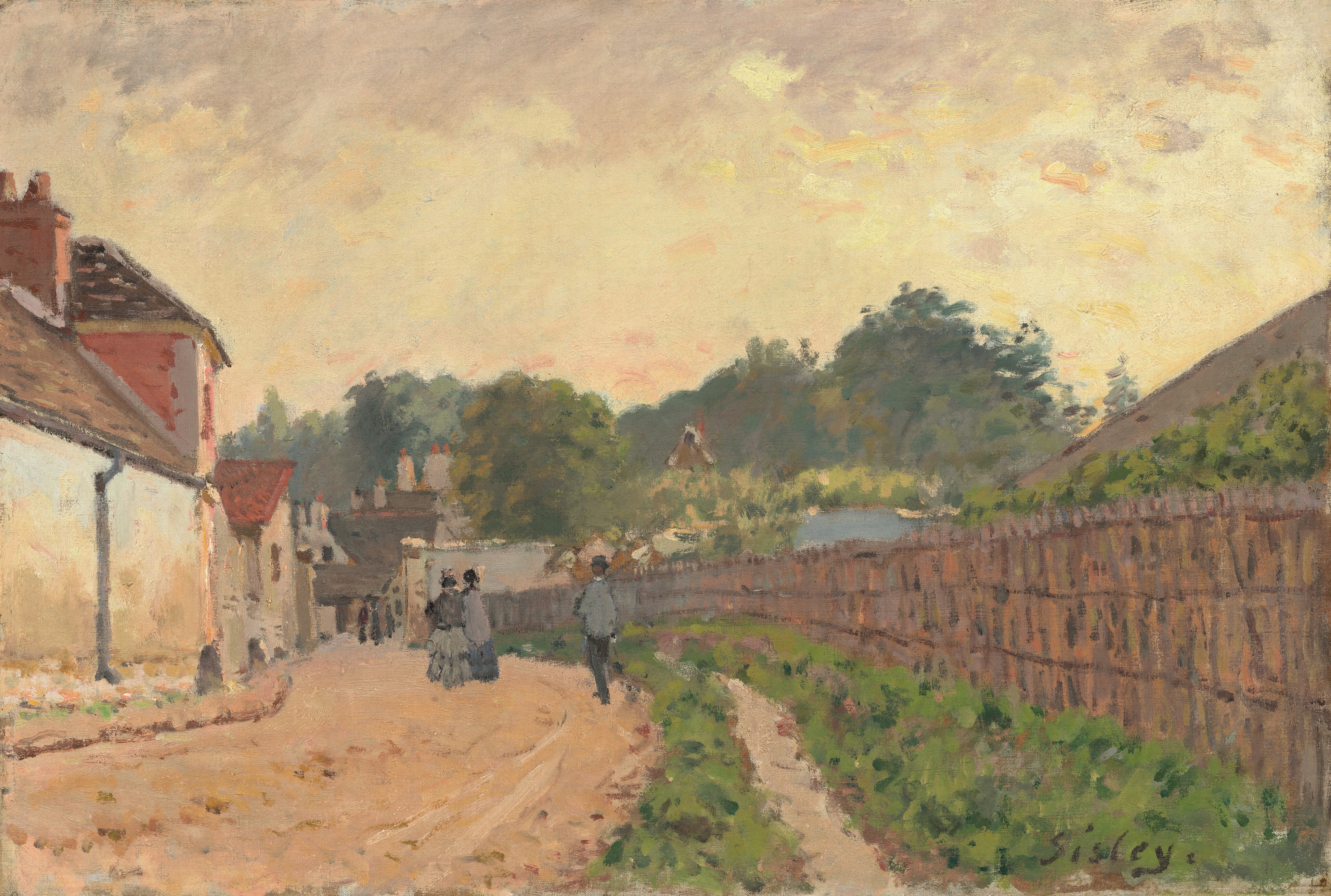This painting depicts a street scene from a slightly elevated perspective. A tan dirt road stretches into the distance, lined with white houses on the left side. On the right side is a tall fence, and behind it the green tops of trees or bushes are visible. People in dark clothing walk along the street in the distance. Beyond them, taller trees are visible, with more buildings below them. The sky above is softly colored in hues of yellow, peach, and gray. The brushstrokes are loose and visible.