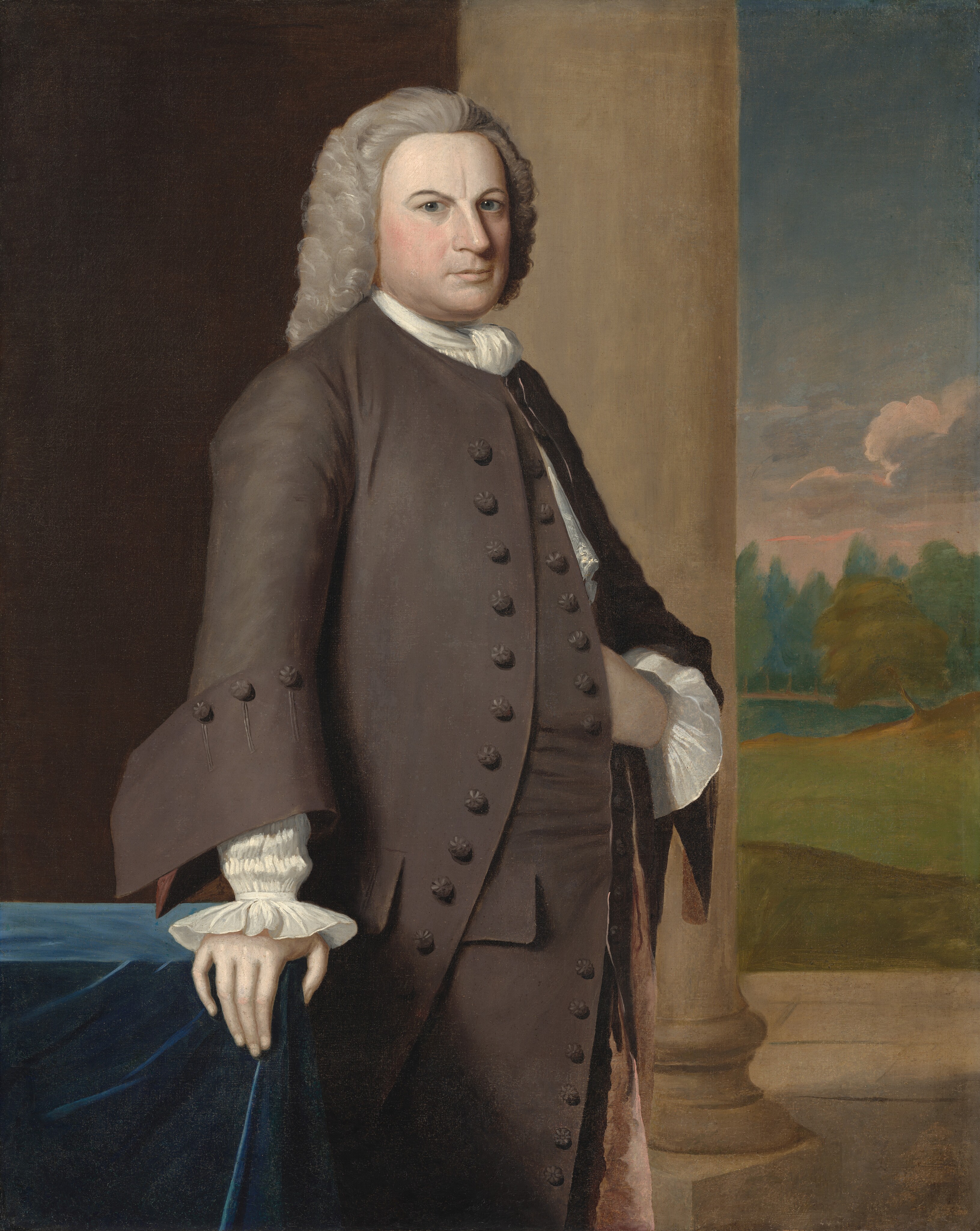 The portrait depicts a man standing with one hand resting on a blue fabric covered table. He has a dignified posture, relaxed shoulders, and a calm facial expression. He has fair skin, a rounded face, grayish-white hair styled in curls typical of the 18th century. The man is dressed in a dark coat with decorative buttons and wide-cuffed sleeves showing white ruffled cuffs. He wears a white cravat. The background includes a stone column and a view of a landscape with a blue sky, hills, clouds, and trees.