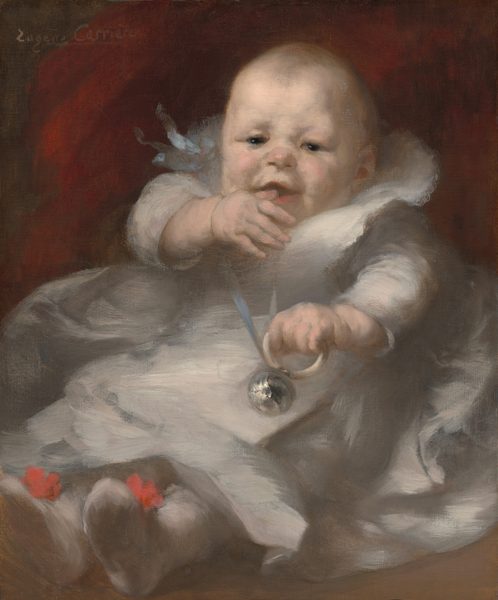 This image shows a young baby from head to toe. The baby is upright, possibly seated or reclined, with soft, rounded features, rosy cheeks, small eyes, and a slight smile. The hair is sparse and light-colored. The baby is dressed in a light-colored garment with red accents on the feet, likely the tops of booties or socks. The baby is holding a silver object attached to a ribbon or string in one hand, possibly a rattle. The background is dark with some red tones, offering a contrast with the light clothing. The artist's name, "Eugène Carrière," is visible in the upper left corner as a signature.