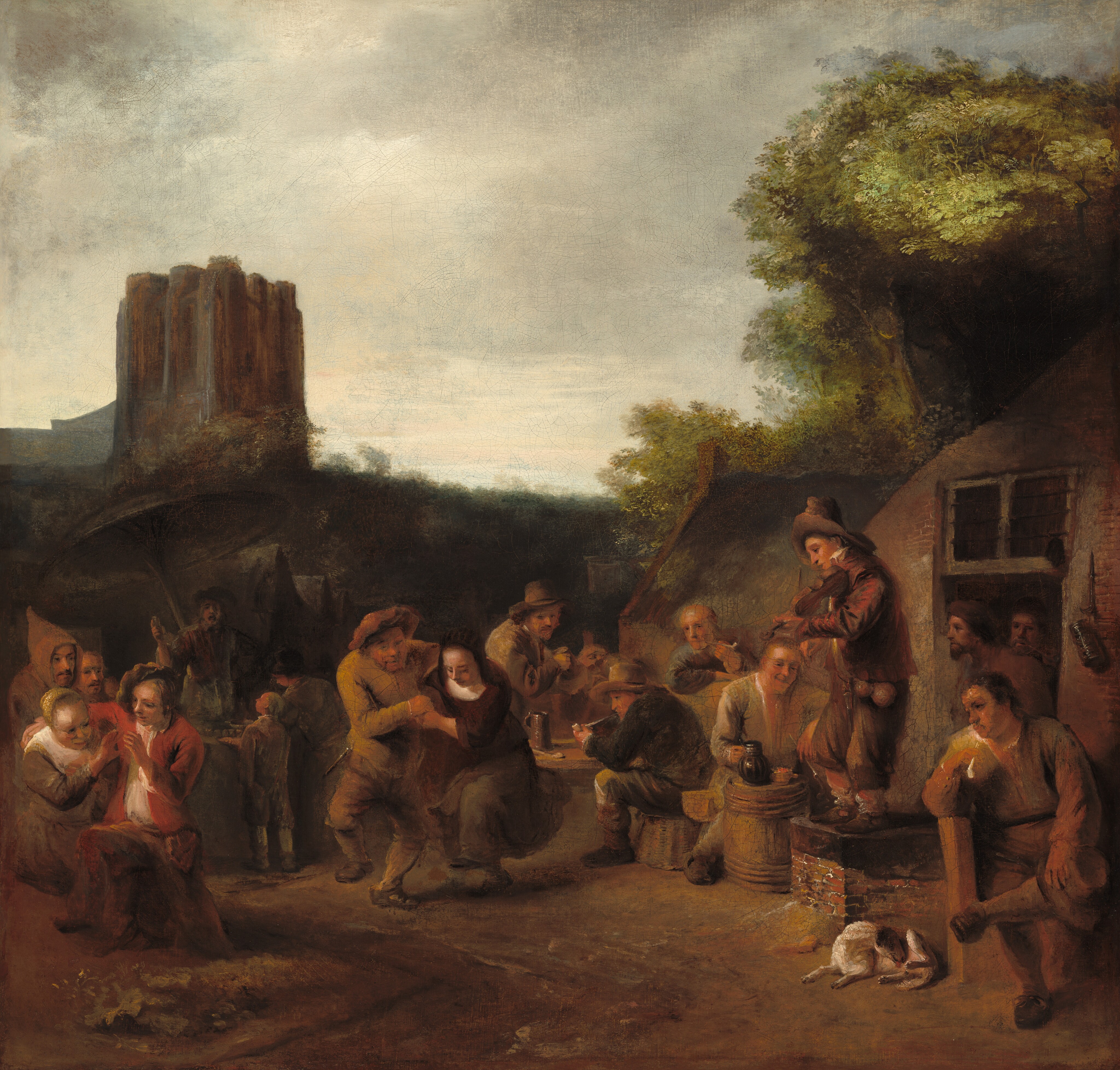 This is a painting of an outdoor scene with numerous people engaged in various activities; some are sitting by low tables, some are dancing, and some are conversing. One person on the right stands on a surface and plays a stringed instrument like a fiddle or violin. The people wear clothes in earth shades of brown, tan, black, and dark red. The background includes a rustic building with a thatched roof, leafy green trees, and what may be a tall castle or church on the left. The cloudy sky above is in shades of gray.