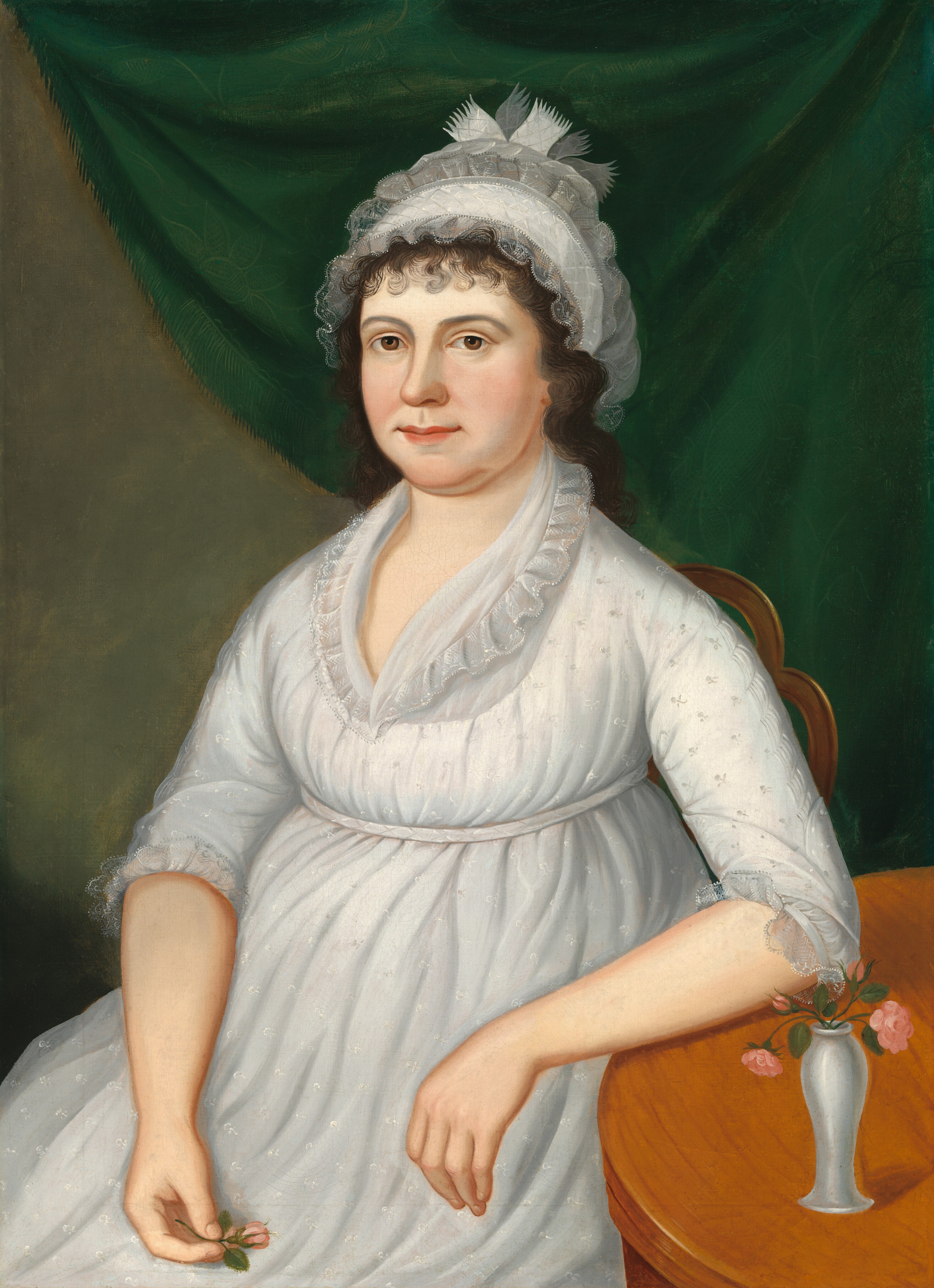 The image is a portrait showing the upper body of a woman seated next to a wooden table. She is positioned facing slightly to the right, with her arm resting on the table and holding a small pink rose in her left hand. Her facial features include a calm expression, rosy cheeks, and visible brown eyes under dark, curly hair that frames her face, partly obscured by a white lace cap with decorative fabric elements. She is wearing a white, high-waisted gown with long sleeves with lace trim and delicate embroidery details. On the table, there is a small white vase with similar pink roses, adding a decorative element. The background consists of a deep green drapery.
