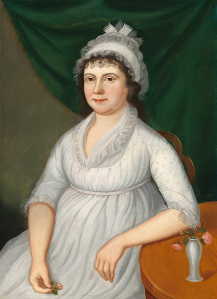 The image is a portrait showing the upper body of a woman seated next to a wooden table. She is positioned facing slightly to the right, with her arm resting on the table and holding a small pink rose in her left hand. Her facial features include a calm expression, rosy cheeks, and visible brown eyes under dark, curly hair that frames her face, partly obscured by a white lace cap with decorative fabric elements. She is wearing a white, high-waisted gown with long sleeves with lace trim and delicate embroidery details. On the table, there is a small white vase with similar pink roses, adding a decorative element. The background consists of a deep green drapery.