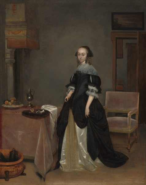 This painting shows a woman standing in a room, her body slightly turned to the left and her head turned to face us. She has a fair complexion, arched dark eyebrows, a pointed nose, a high forehead, and curly hair in an updo under a short black headdress. She is wearing a black gown with white lace trim on the collar and sleeves and a cream-colored underskirt with gold embroidery. Her hands are in her skirt, holding bunches of black fabric. She stands next to a table on the left with various items on it, including a bowl of fruit, a silver pitcher, and a black tray. A chair is positioned behind her to the right, and a partially open door is visible in the background. The wall behind her is pale gray-blue, and the ground is tan.