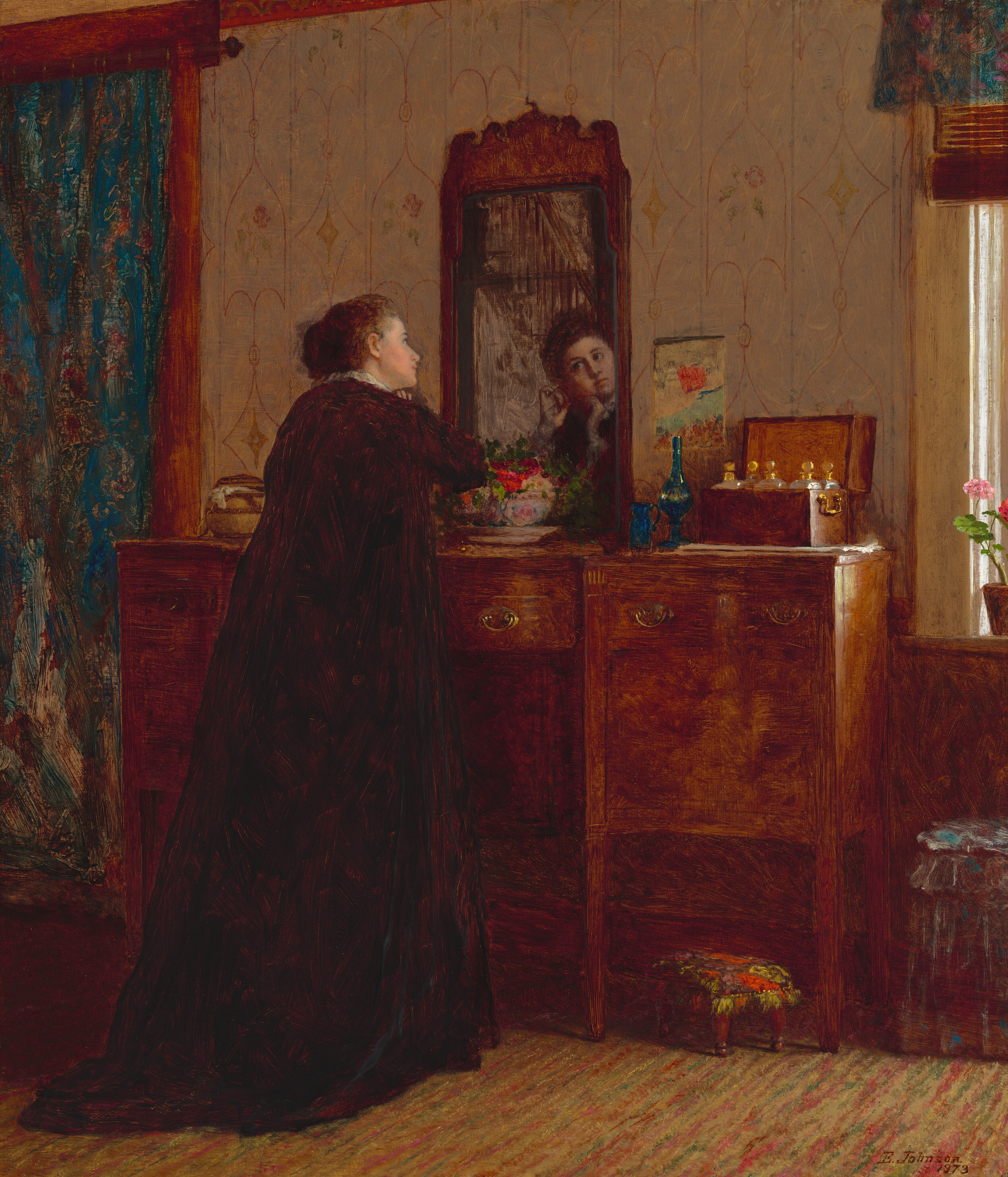 This painting shows a woman standing in front of a wooden dresser with a mirror, her back to us. We can see her face reflected in the mirror. She is gazing at her reflection with both of her hands lifted towards her slightly tilted face. The woman has pale skin and dark hair neatly styled away from her face, and she is wearing a dark gown with white ruffles around the neck. On the dresser, there is a floral arrangement, a pair of blue bottles, and a wooden box holding more containers. Behind the dresser, the background includes patterned wallpaper, a blue curtain on the left, and sunlight coming through a window on the right. The floor appears to be covered by a patterned rug.