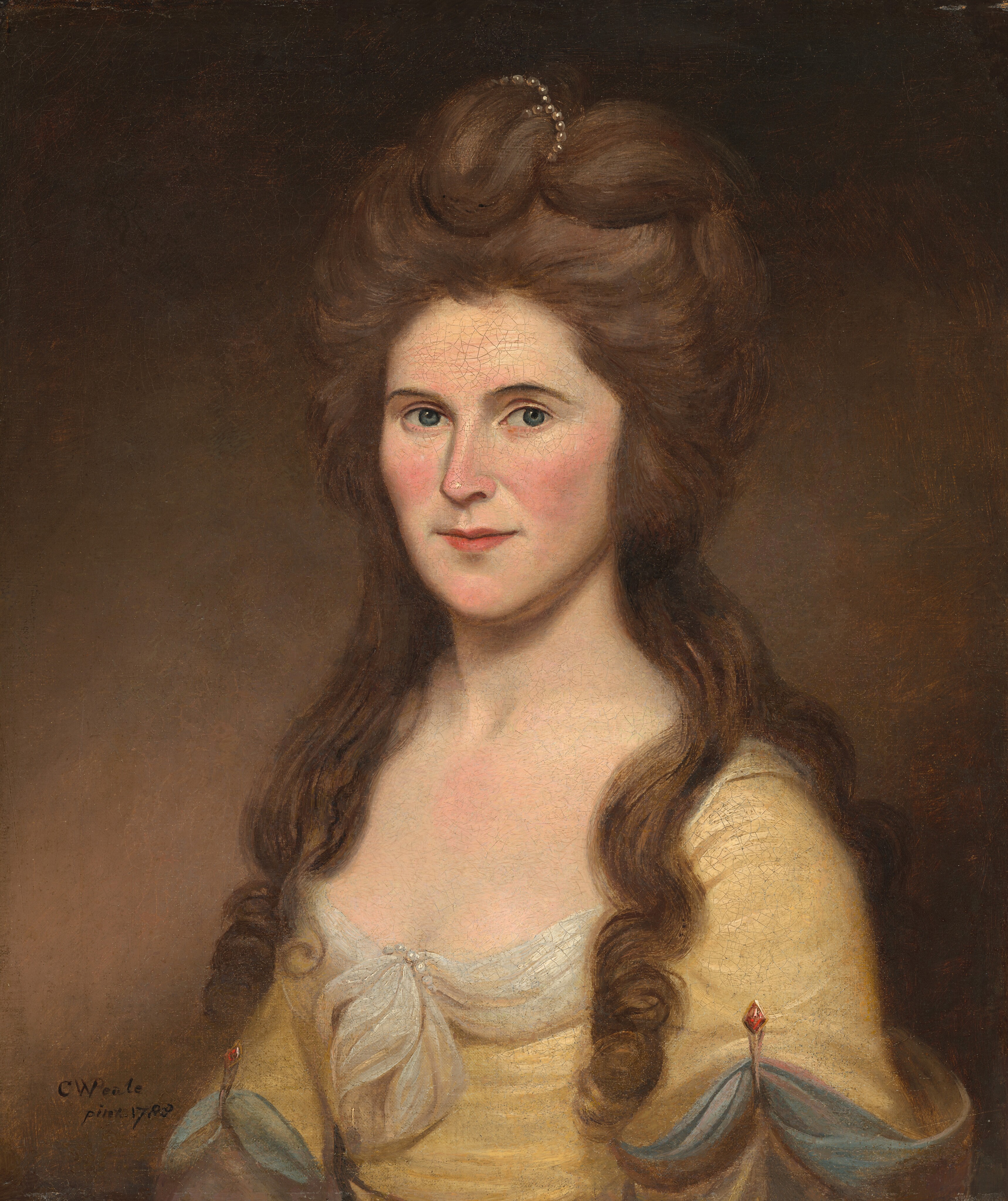 The painting shows a woman's figure from the waist up, seated, and her body is turned slightly to our left. Her head is turned to face the viewer. Her facial features are delicate, with a fair complexion, soft pink cheeks, and light blue eyes. She has long brown hair styled in voluminous waves, pulled back halfway and adorned with pearls. She is wearing a pale yellow gown with a low neckline accented by lace edges and a fabric decoration at the center of the bust. Her sleeves feature blue ribbon details and red jewel embellishments at the shoulders. The background is a muted brown.