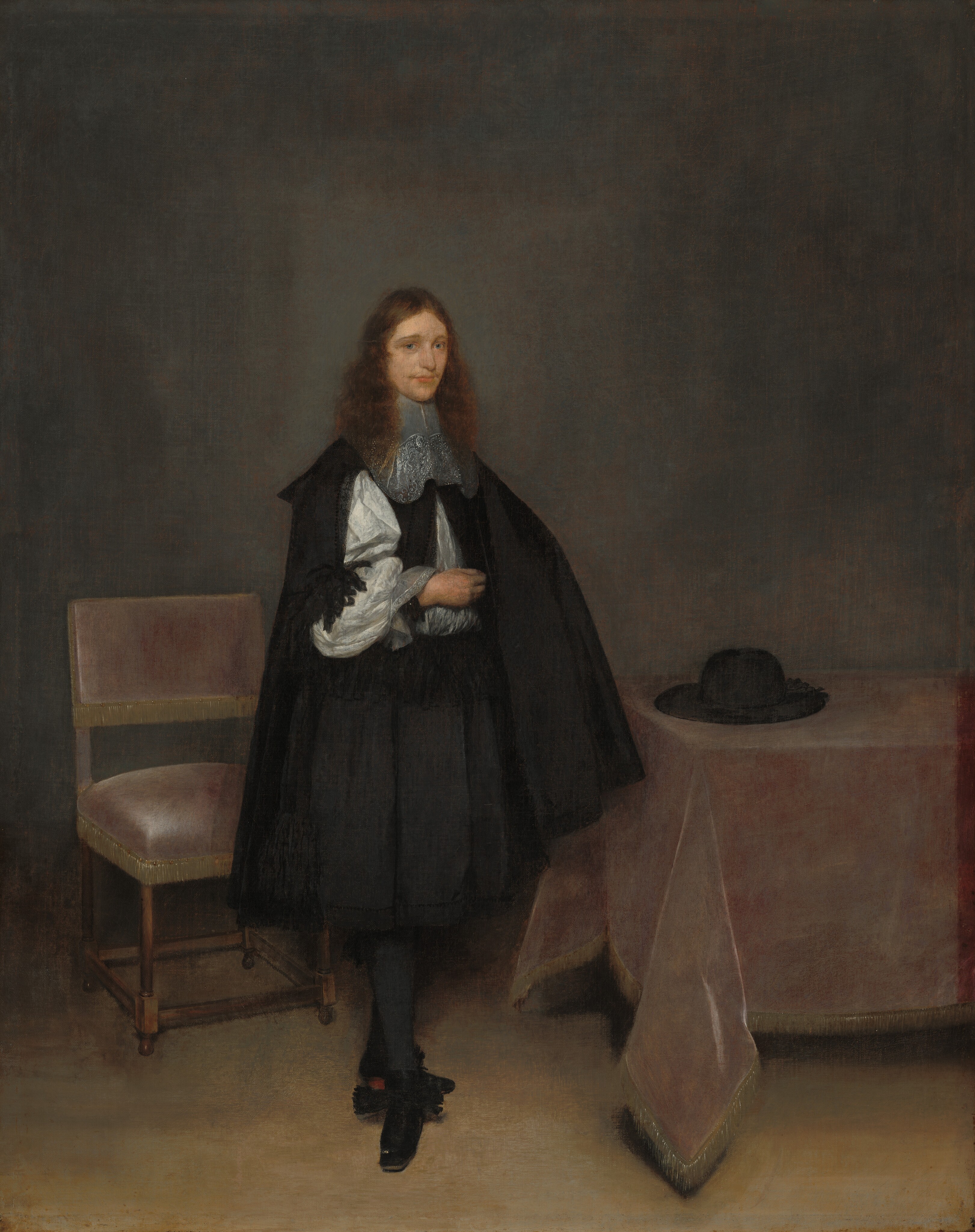 This painting depicts a person standing indoors, slightly turned to the right. They have pale skin, long, curly, reddish-brown hair, a faint mustache, blue eyes, and pink lips in a neutral expression. Their right hand is touching their chest, and their other hand is hidden under their cloak. Their legs are slightly crossed. The person wears a dark cape over a black doublet, a white shirt with ruffled cuffs, an ornate lacy collar, a knee-length black skirt, black stockings, and black shoes with ruffles. In the background, there is a chair on the left with a pale-pink cushion, a table on the right with a pale-pink tablecloth and a black hat on top of it, and a plain gray wall in the background.