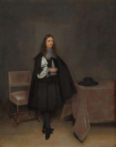 This painting depicts a person standing indoors, slightly turned to the right. They have pale skin, long, curly, reddish-brown hair, a faint mustache, blue eyes, and pink lips in a neutral expression. Their right hand is touching their chest, and their other hand is hidden under their cloak. Their legs are slightly crossed. The person wears a dark cape over a black doublet, a white shirt with ruffled cuffs, an ornate lacy collar, a knee-length black skirt, black stockings, and black shoes with ruffles. In the background, there is a chair on the left with a pale-pink cushion, a table on the right with a pale-pink tablecloth and a black hat on top of it, and a plain gray wall in the background.