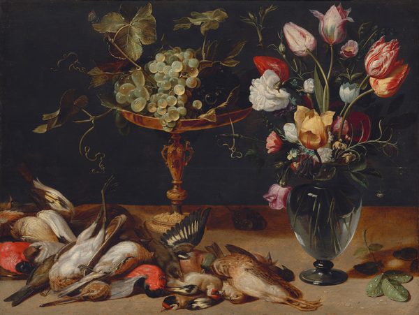 In the center of this painting there is a large ornate golden goblet overflowing with grapes and green leaves. On the right, a dark glass vase is filled with various colorful flowers, such as tulips and roses in shades of yellow, pink, white and red. Below the goblet, in the bottom left corner of the painting, is a grouping of several birds. The birds are laying on top of one another in a large pile, some upside down and with legs or wings extended. Some of the birds have colorful plumage in shades of red or green. The goblet, vase, and birds rest on a brown surface, and the wall behind the objects is black.