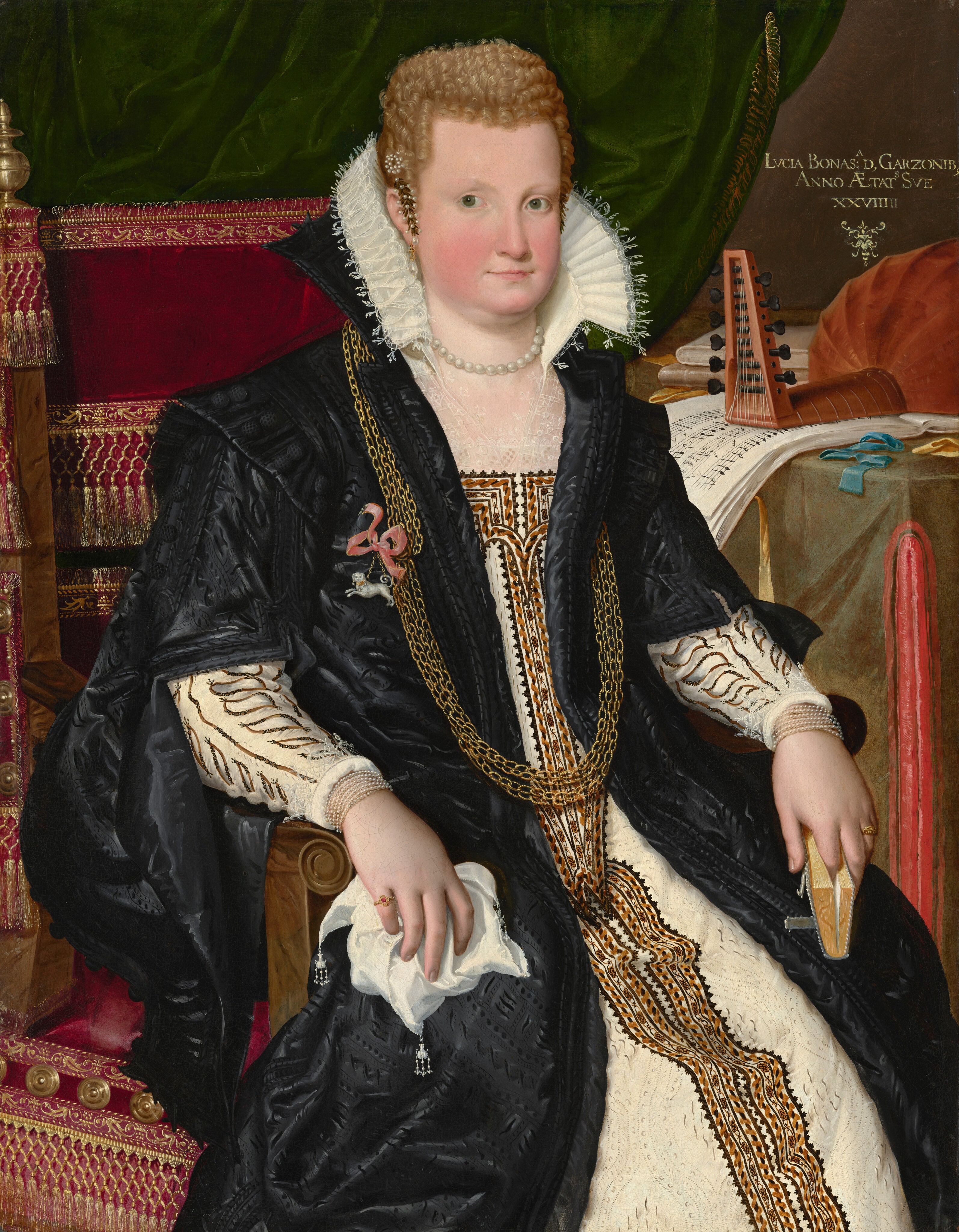 This painting is a portrait of a seated woman, her body turned slightly to the right and her head looking towards us. She has a fair complexion, with round, rosy cheeks, pale eyebrows, large green eyes, and thin pink lips in a neutral expression. Her light blonde hair is styled in short curls close to her head and adorned with pearls and a clip or barrette in the shape of small leaves. She is dressed in a cream-colored dress with intricate embroidery in shades of brown, gold, and black. On top of the dress is a long black garment that flows over her shoulders and upper arms and has a high collar, decorative buttons, and more embroidery.  Her gown is paired with a ruffled white collar and lace at her neckline and wrists. Long gold chains drape across her chest, and she wears a pearl necklace around her neck and two small golden rings on her fingers. In her left hand, our right, she holds a small book, while a lace handkerchief is in her right hand, our left. She wears pearl earrings, strands of pearl bracelets, and a decorative brooch in the shape of a white dog with a pink bow. The seat she sits on is wooden with dark red cushions decorated with golden tassels and embroidery, behind her on the left is a draped green cloth, and on the right, a table is covered with music sheets, a stringed instrument, books, and small blue and yellow ribbons. The wall behind the table is brown, and there is some gold lettering in the top right corner.