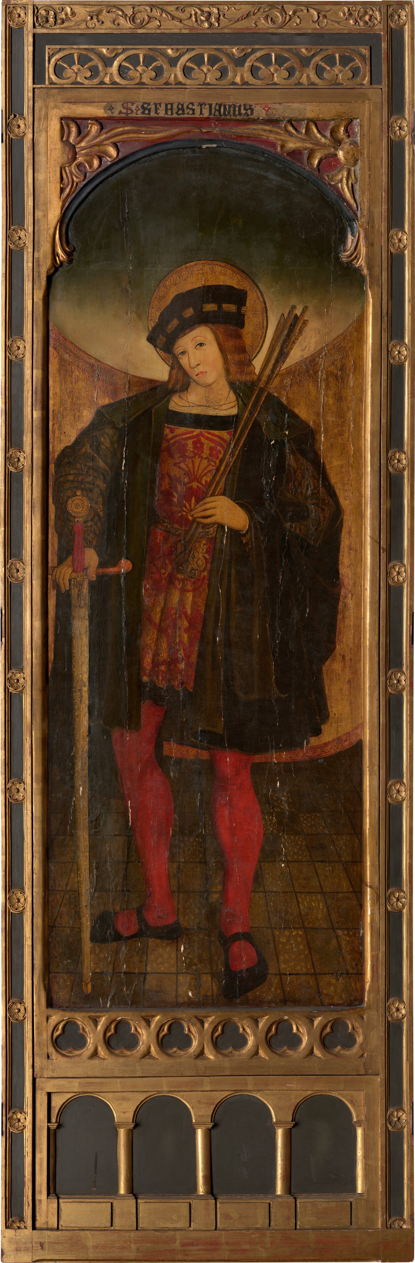 The image shows a full-length painting of a person standing. The figure is positioned in a slightly relaxed stance, holding a staff or arrows in one hand and a sword with an ornate handle in the other. The person's face is depicted with soft, youthful features and long, flowing hair, covered with a hat or headpiece. The clothing consists of a long, dark cloak or robe layered over a richly decorated red garment and red tights accompanied by black shoes. A halo is observed behind the person's head. The painting is framed in an ornate wooden structure with intricate carvings and gold detailing, with the name "S. SEBASTIANUS" inscribed at the top. The background features a tiled floor with a golden-brown gradient backdrop.
