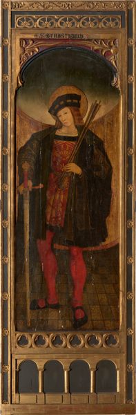 The image shows a full-length painting of a person standing. The figure is positioned in a slightly relaxed stance, holding a staff or arrows in one hand and a sword with an ornate handle in the other. The person's face is depicted with soft, youthful features and long, flowing hair, covered with a hat or headpiece. The clothing consists of a long, dark cloak or robe layered over a richly decorated red garment and red tights accompanied by black shoes. A halo is observed behind the person's head. The painting is framed in an ornate wooden structure with intricate carvings and gold detailing, with the name "S. SEBASTIANUS" inscribed at the top. The background features a tiled floor with a golden-brown gradient backdrop.