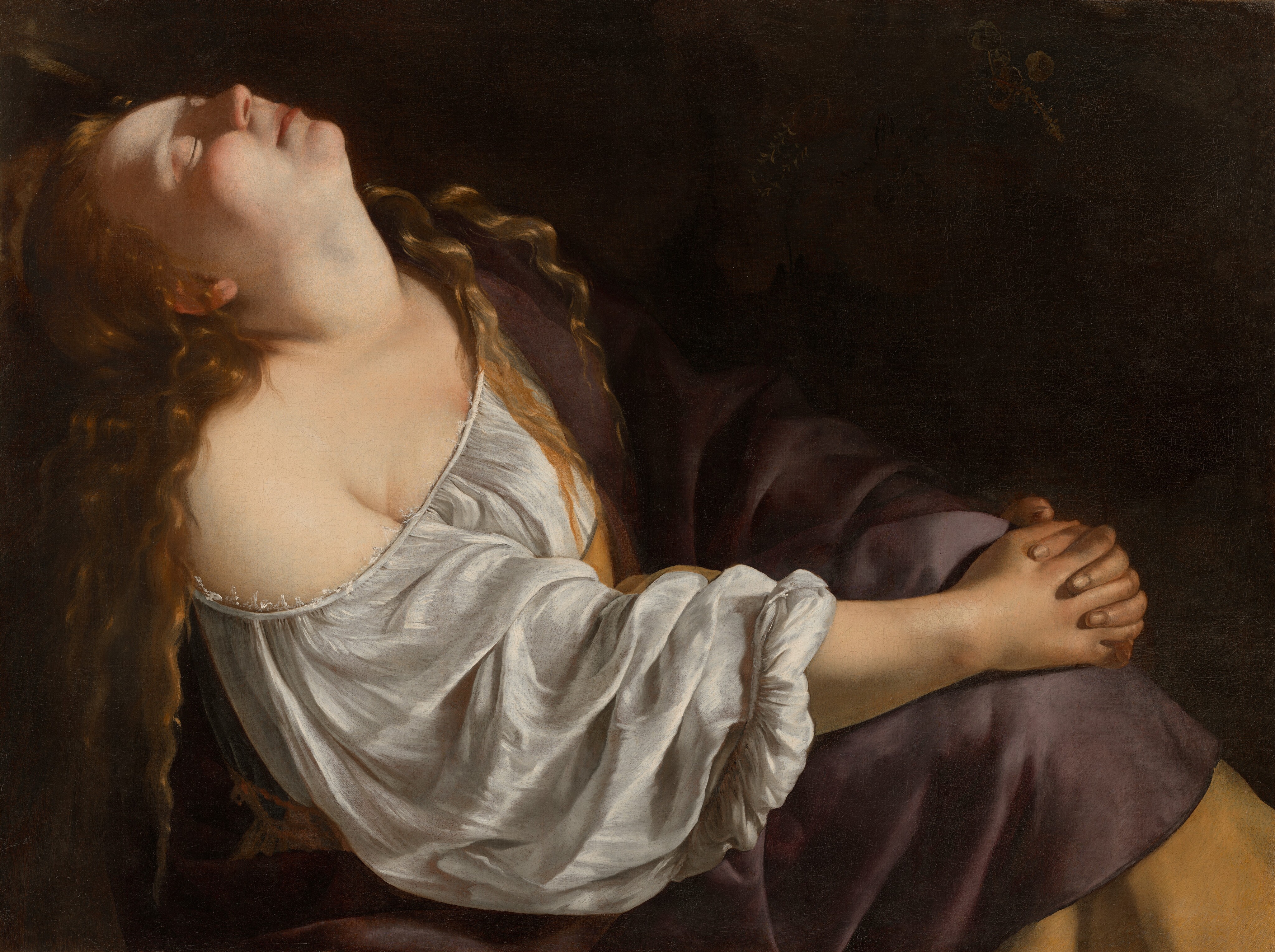 This painting shows a woman sitting with her body turned to the right, her hands clasped around her knees and her head tilted up to the ceiling. Her eyes are closed, and her long, wavy hair flows over her shoulders and down her back. She has pale skin, and her face is illuminated by dramatic lighting, highlighting her round cheeks and small nose. She is wearing a white blouse with ruffled sleeves that drapes off of her right shoulder, exposing her neck, sternum, and part of her shoulder. Over this shirt she seems to be wearing a draped purple garment with a yellow border that covers her other shoulder and her legs. The background is dark.