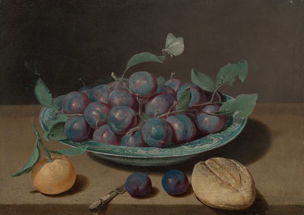 The image depicts a grouping of plums on a ceramic plate, an orange fruit on the left, a loaf of bread on the right, set on a wooden surface against a dark background. The plum colors and green leaves contrast with the orange fruit. The plate has a light design. A white butterfly hovers over the plums.