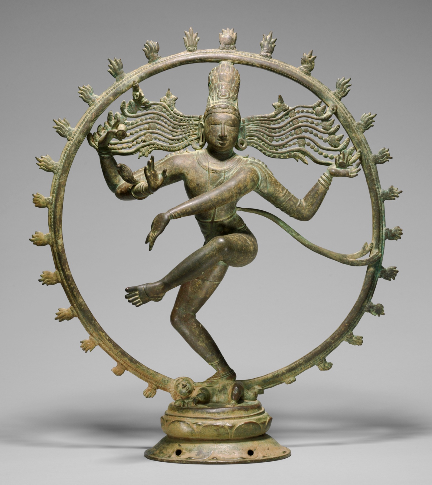 A bronze sculpture of the Hindu divinity Shiva. Shiva, who has two pairs of arms, is depicted dancing inside of a circular ring. Balancing on one leg, Shiva gestures his arms to the side and front, as his loose locks of hair flow behind him.