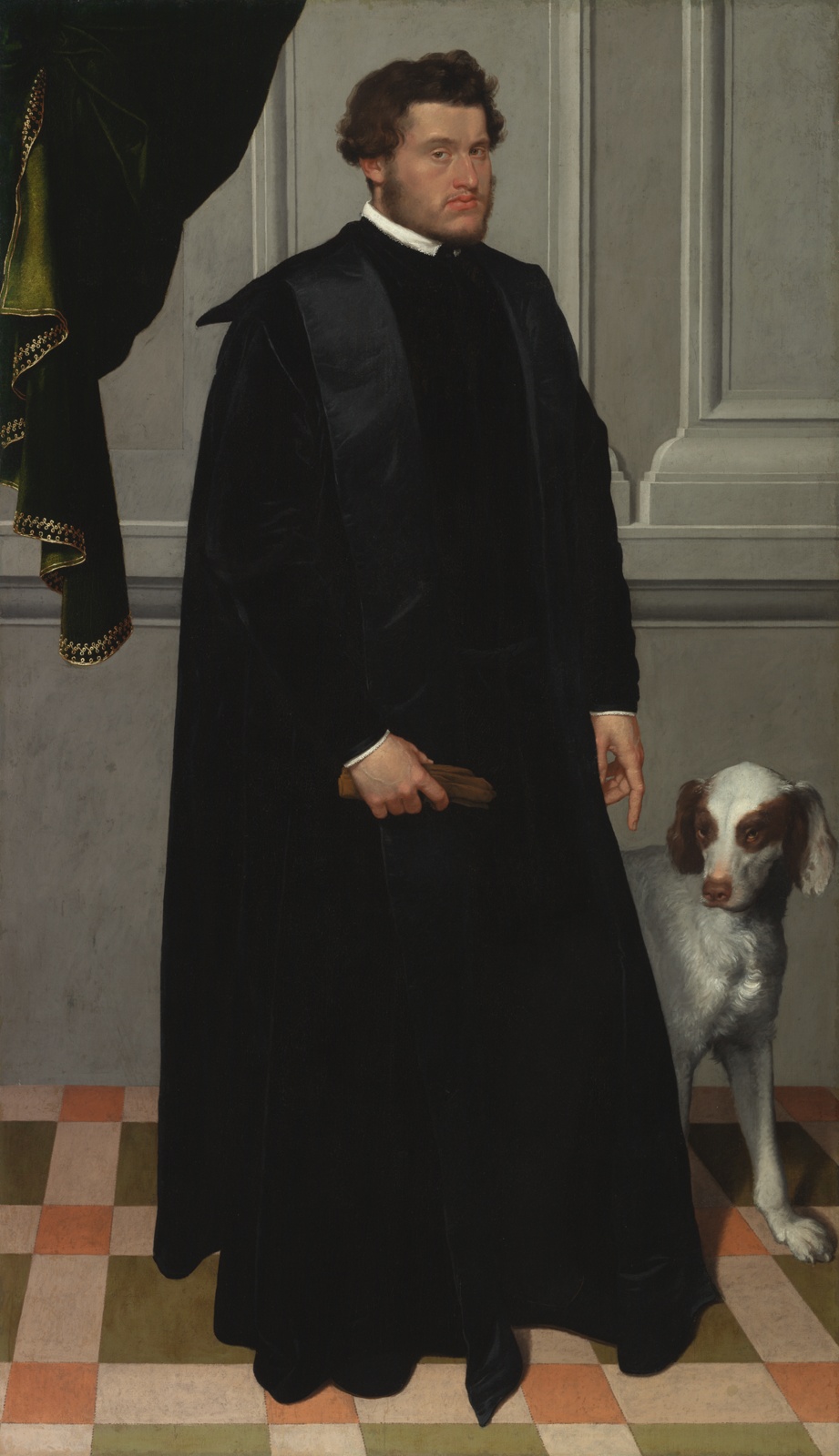 A man with sparse brown facial hair stands in a full-length black robe on a multicolored tiled floor, a white and brown dog behind him.