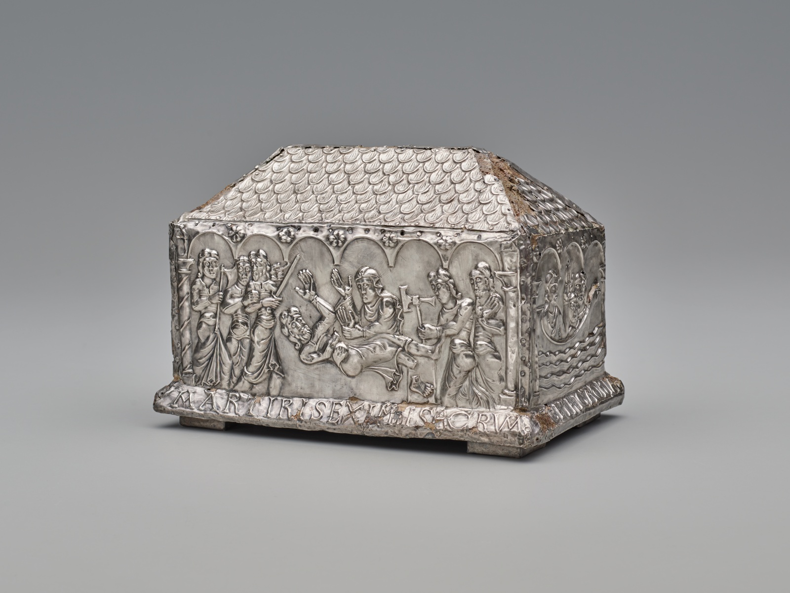 Silver-toned reliquary in the shape of a long rectangle with an upper resembling a pitched roof. In relief on its side, armed figures surround a falling figure in distress as a single figure attacks him.