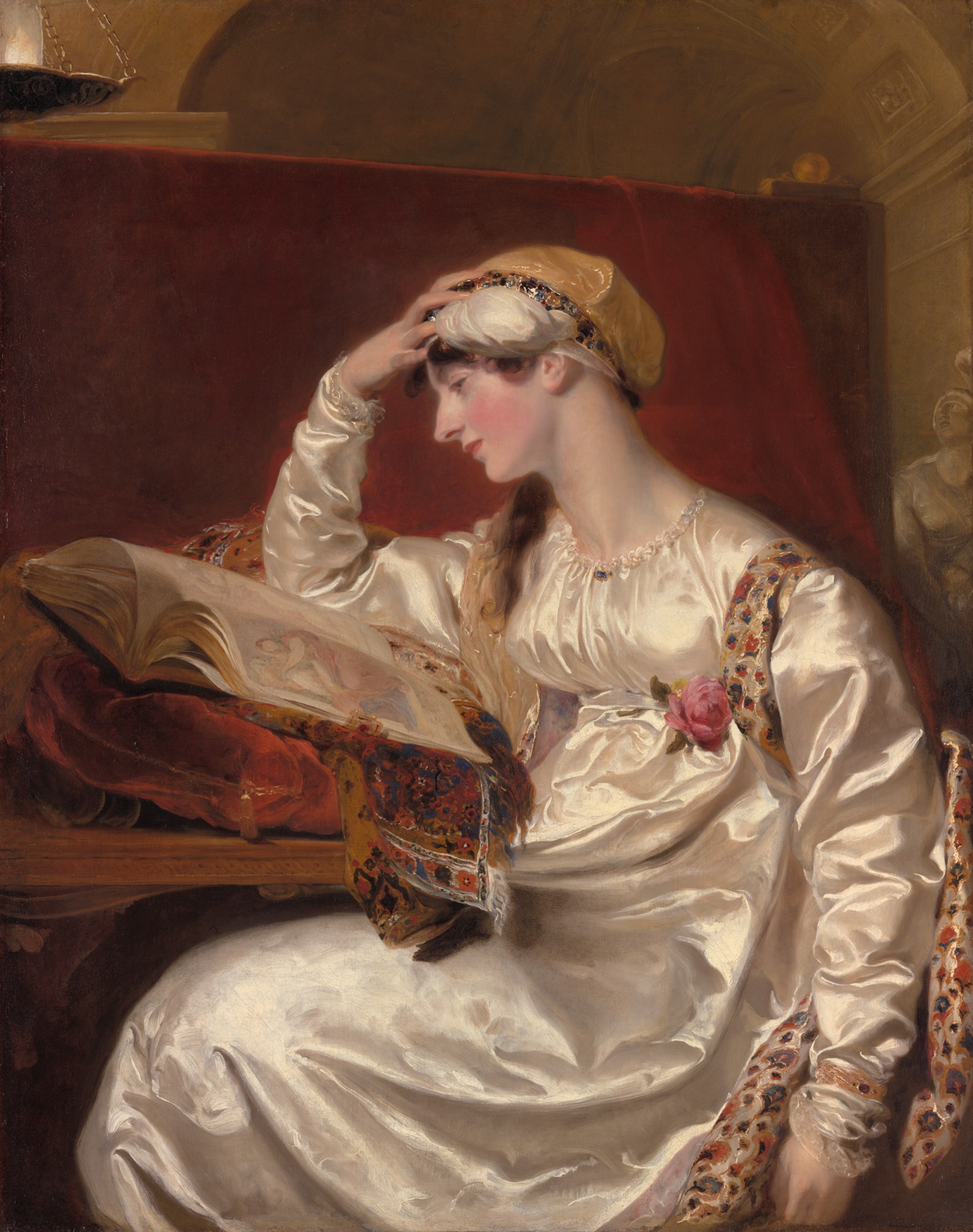 A light-skinned woman in a long, shiny white dress sits at a desk reading a book. The book is propped on a large red pillow. Her head is propped in her hand.