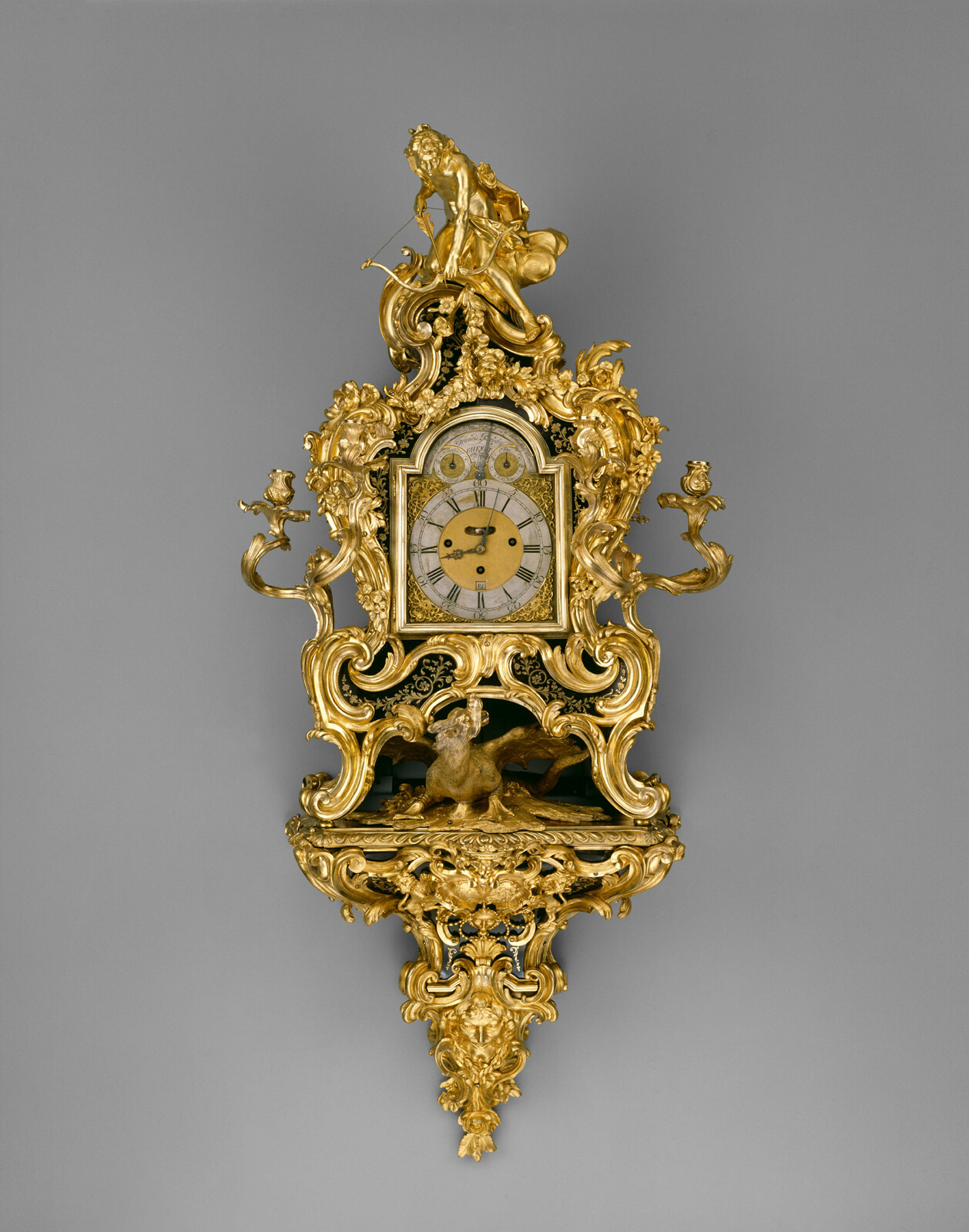 Golden-colored, ornate wall clock adorned with golden flourishes, a bent figure with an arrow at top.