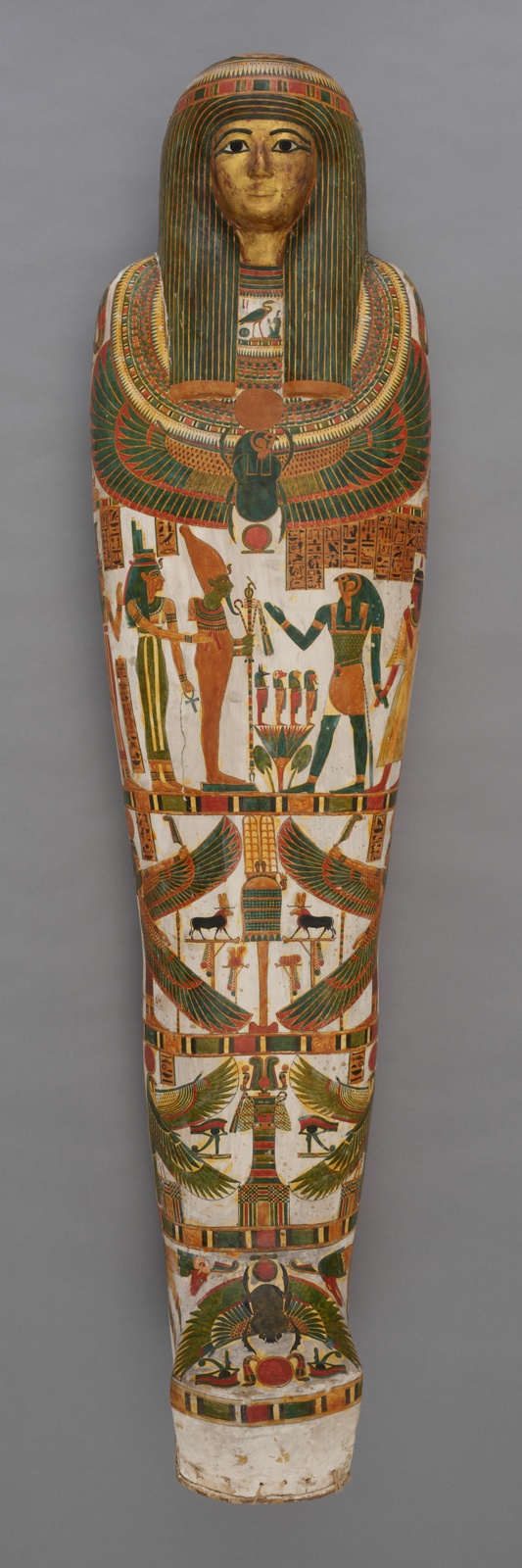 The outer hard case of an Egyptian mummy is painted with a gold face, a blue geometric wig, a colorful bird across the chest, and various scenes across the lower portion featuring figures of gods and goddesses.
