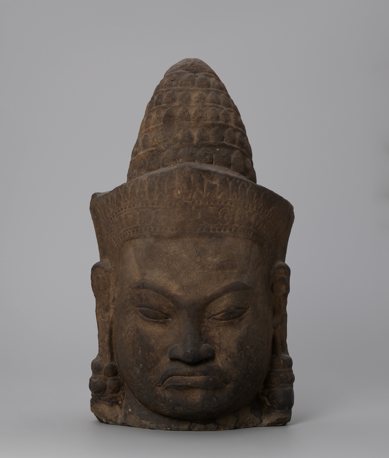 Tan stone carving of a head, features weathered darker, headwear with geometric pattern.